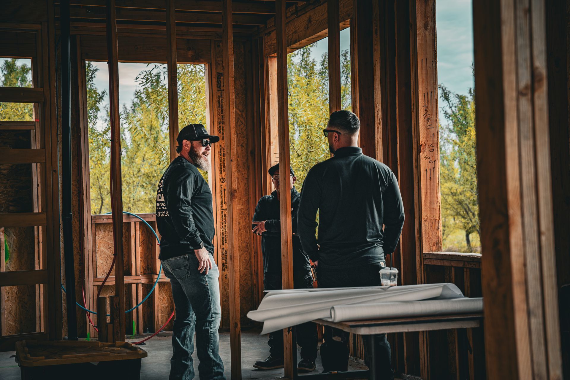 Three workers stand in an unfinished wooden building with large windows and construction materials.