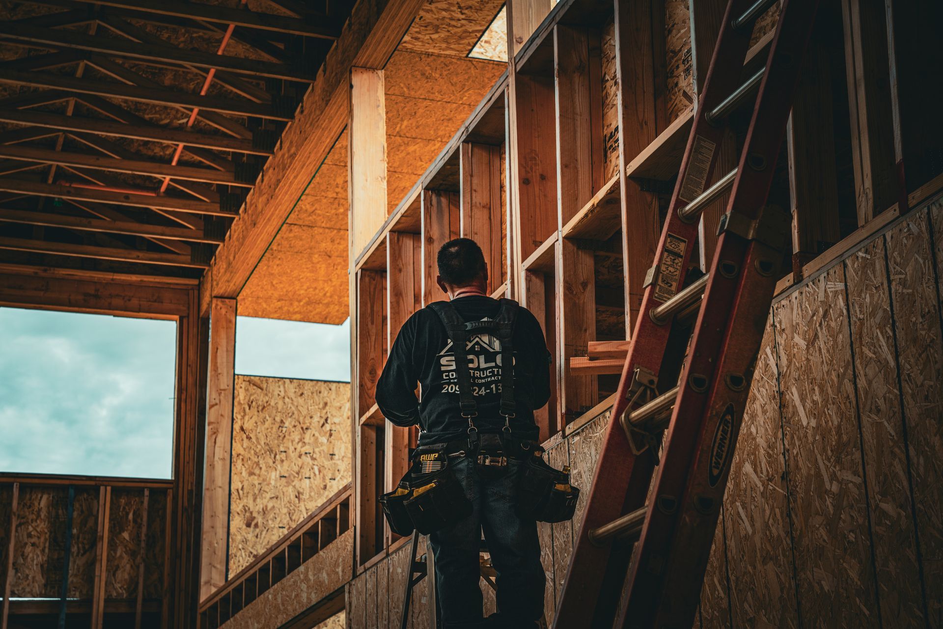 Person climbing a ladder inside a wood-framed building under construction, with warm light and open roof beams