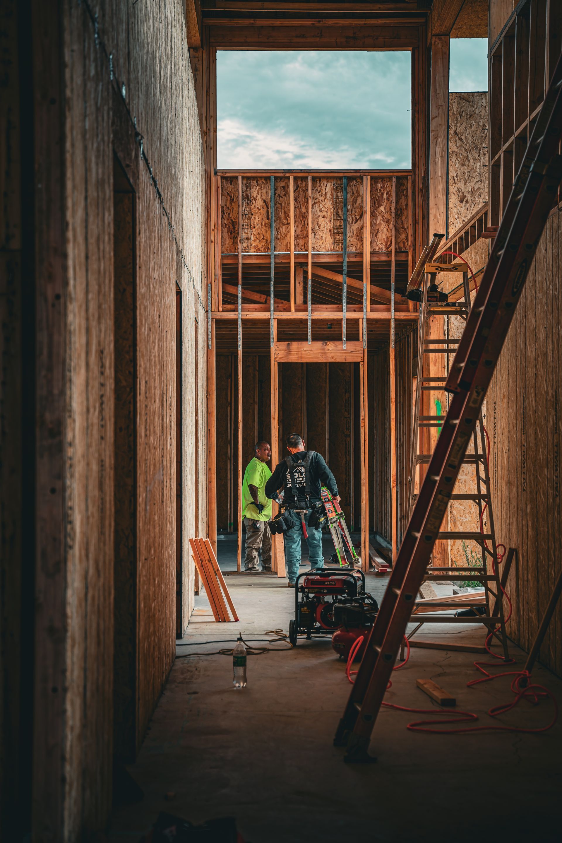 Construction workers inside a framed building interior with ladders and tools, viewed down a narrow hallway