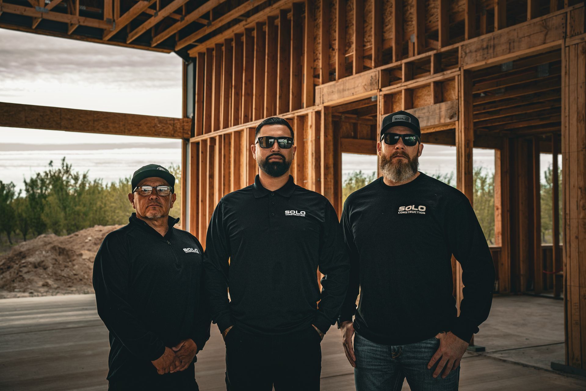 Three men in dark clothing stand inside a wooden house frame under construction.