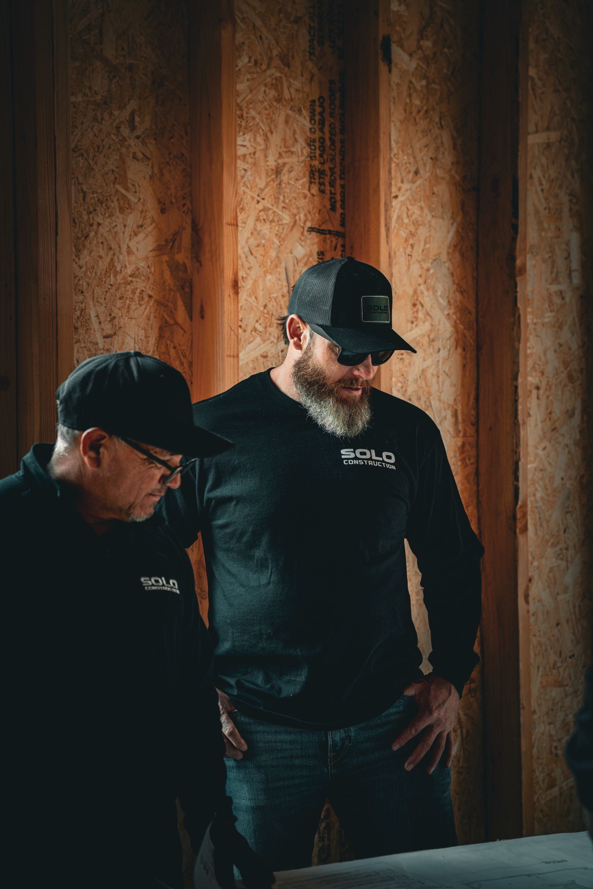 Two men in black shirts and caps standing against a warm wooden wall