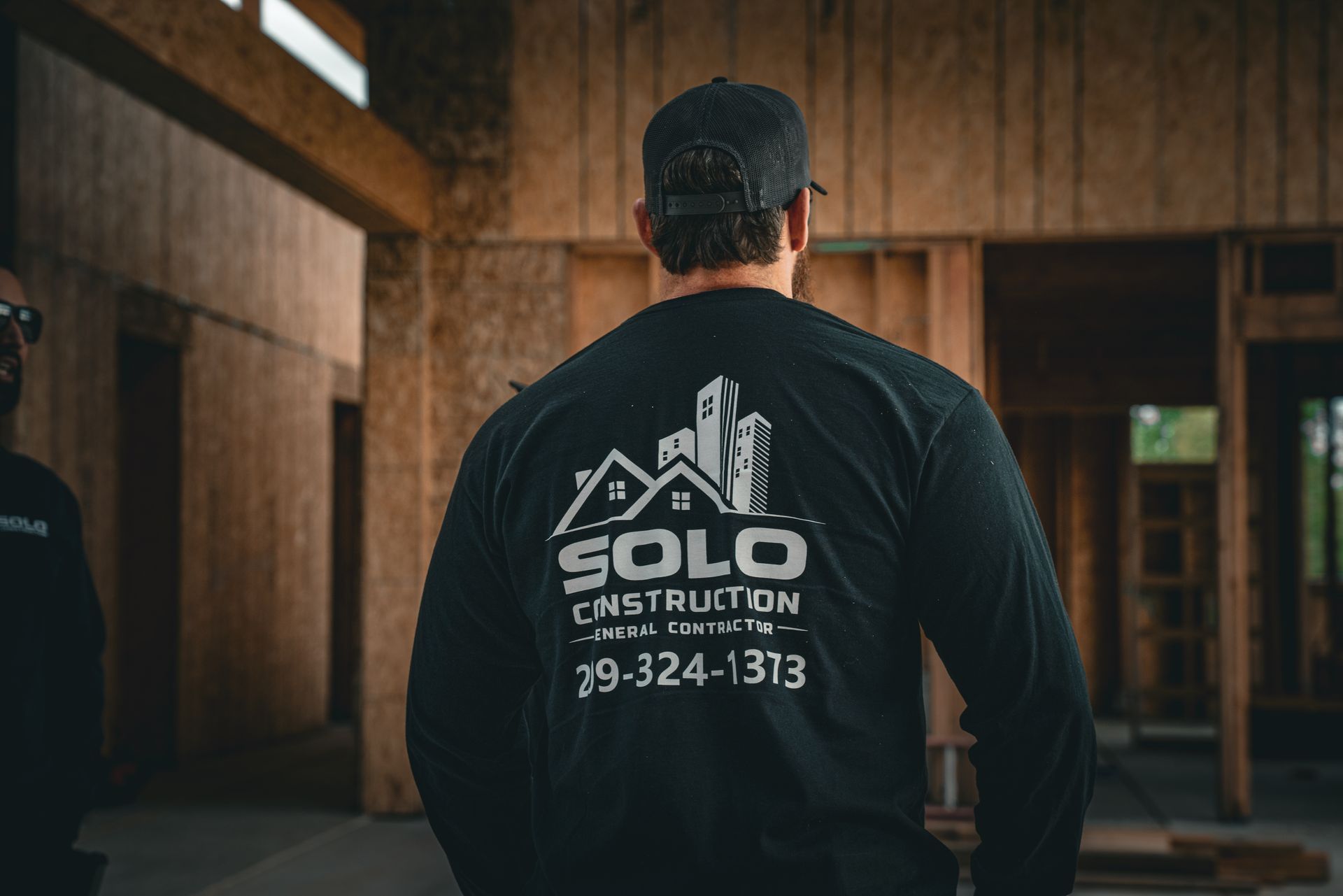 Back view of a person in a black SOLO Construction shirt inside a wood-framed building, with logo and phone number.