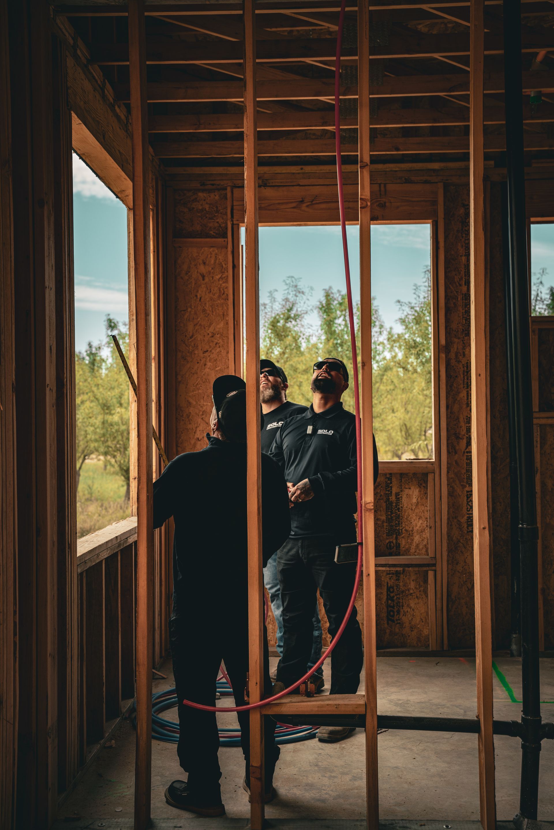 Three people in dark clothing inspect a wooden room under construction with open window framing trees.