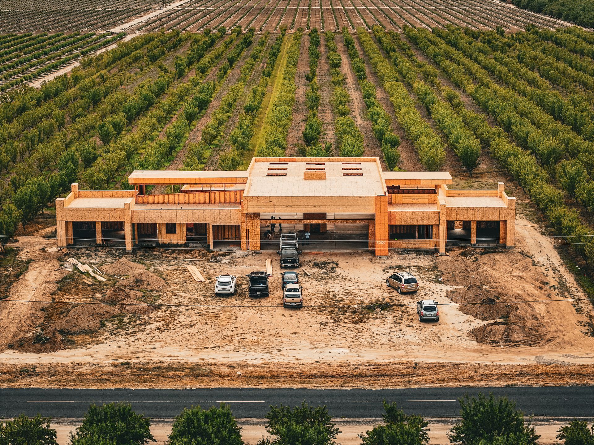 Aerial view of a large orange building under construction beside a road, surrounded by orchards and dirt lots