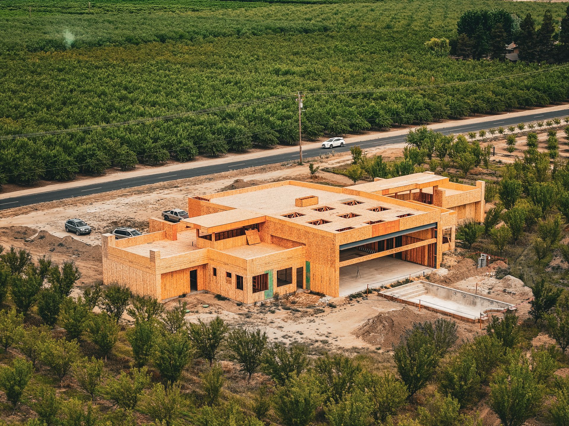 Aerial view of a wooden house under construction beside a road, surrounded by farmland and trees.