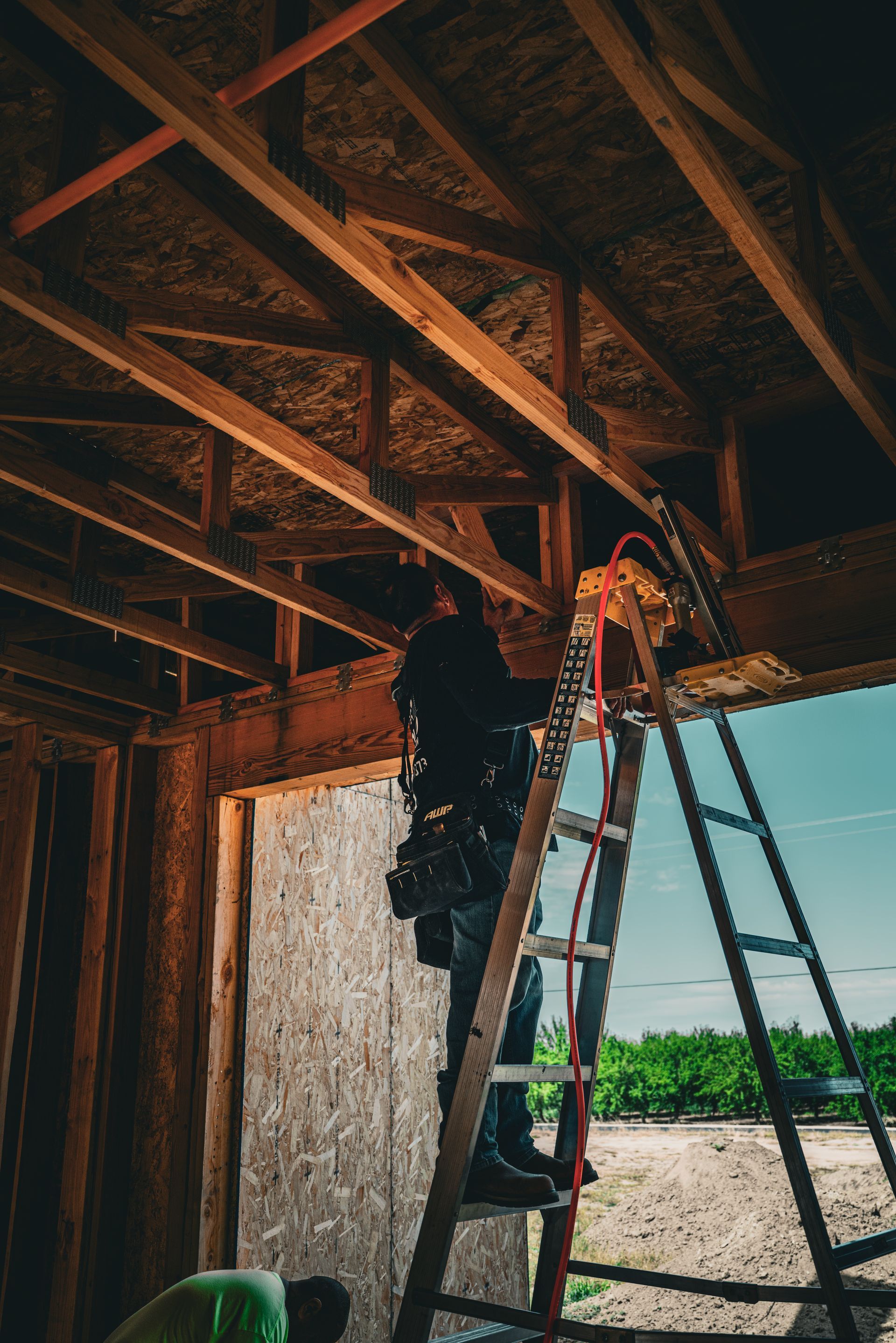 Two workers on a ladder installing wiring under exposed wooden floor joists indoors
