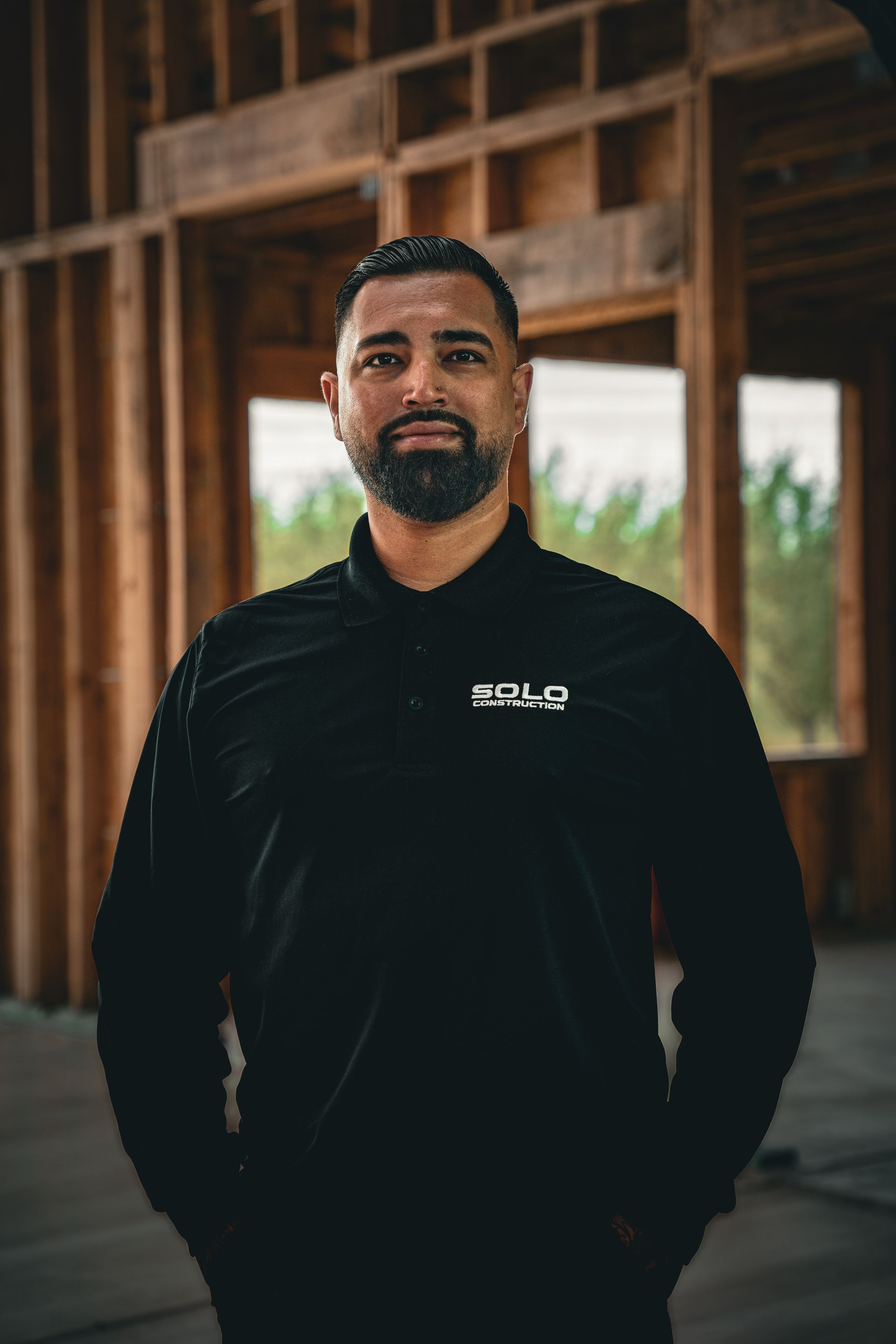 Man standing in a wooden structure, wearing a black shirt, with hands in pockets.