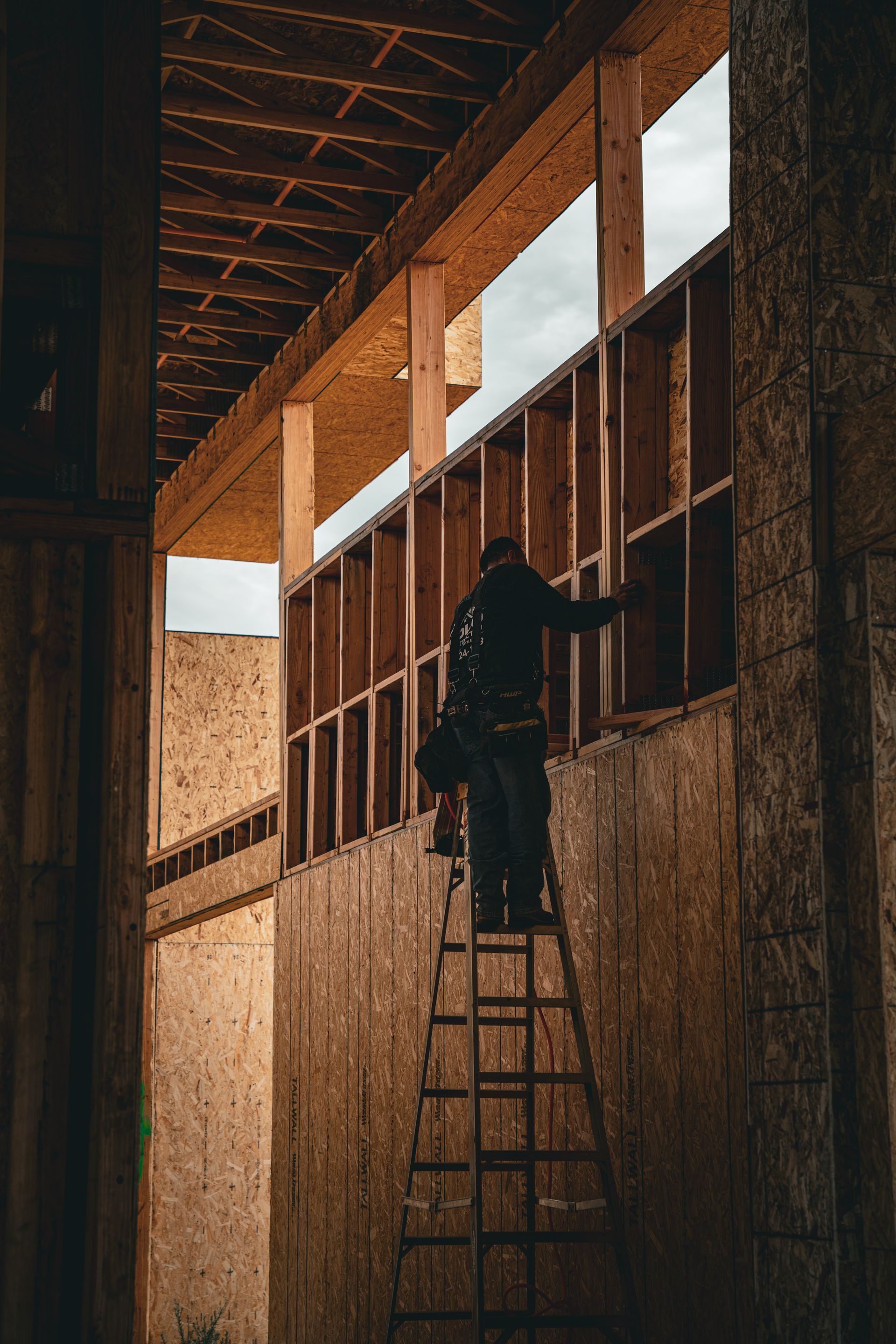 Worker on a ladder installing windows in a narrow, unfinished building corridor, backlit by warm sunlight