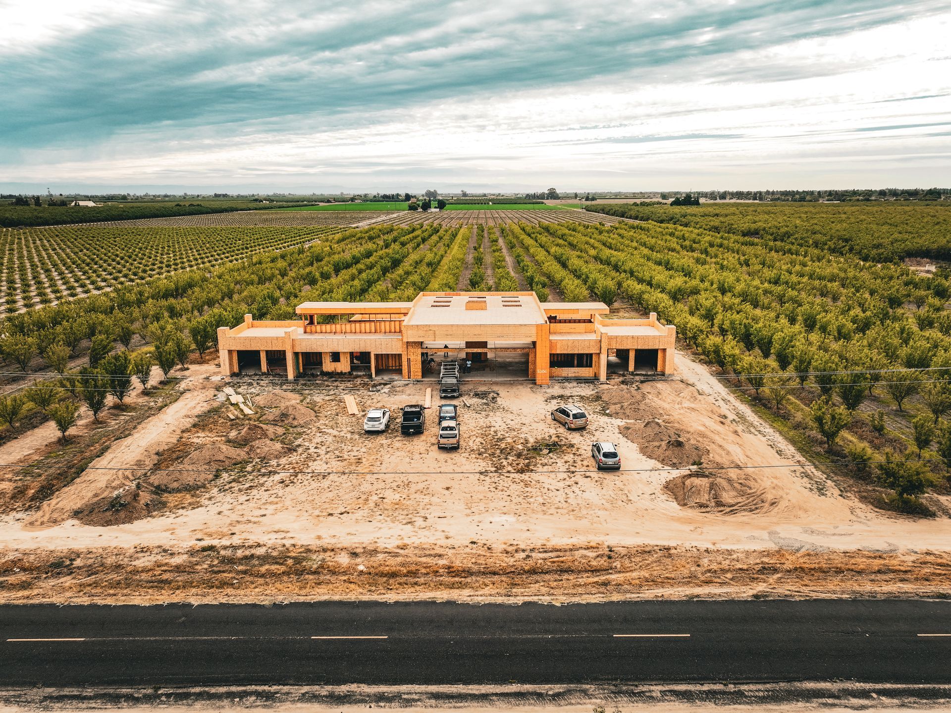 Aerial view of a tan building beside a dirt lot and road, surrounded by rows of orchard trees under a cloudy sky