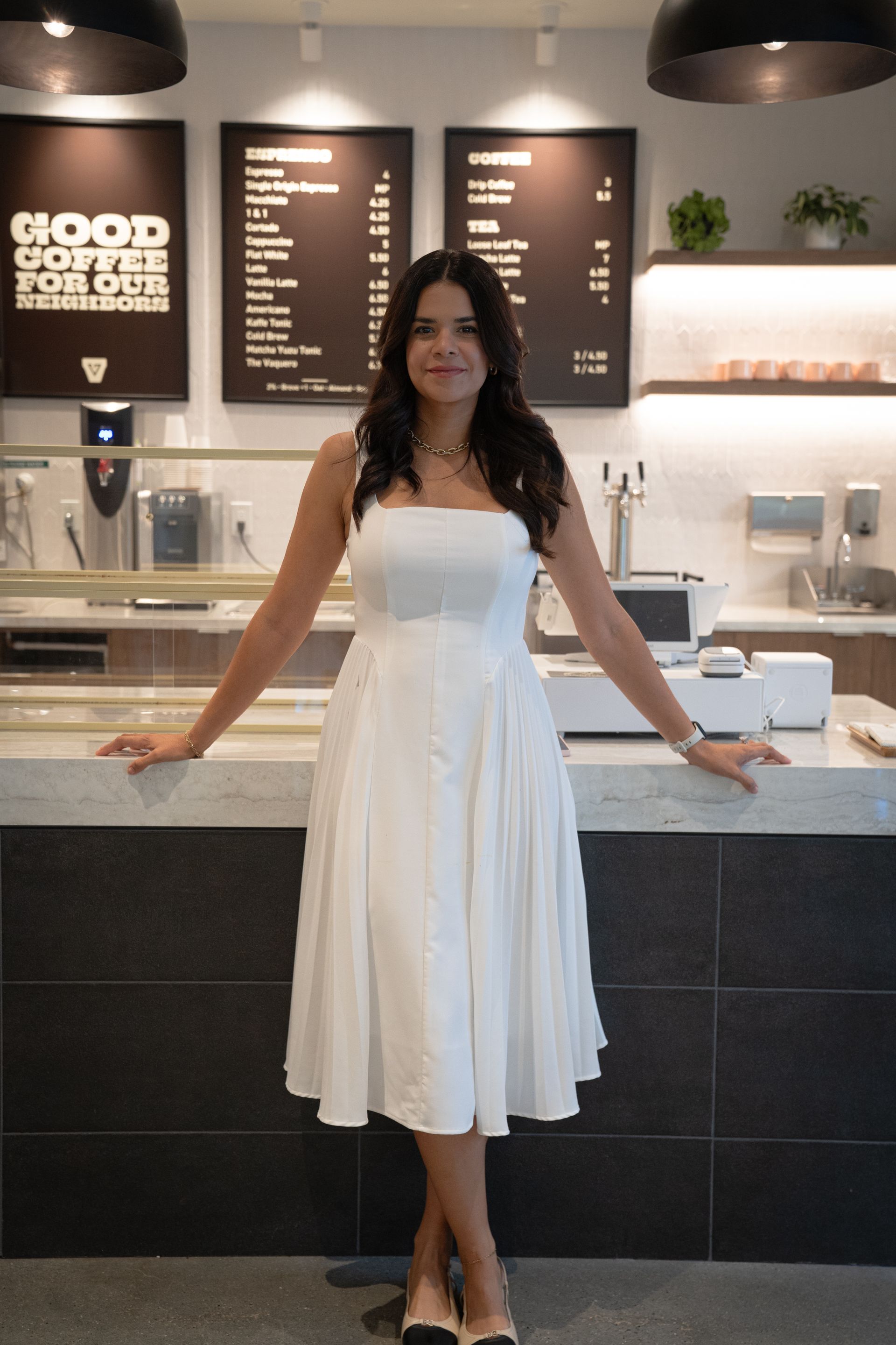 Woman in a white dress standing at a café counter with menu boards and pendant lights behind her.