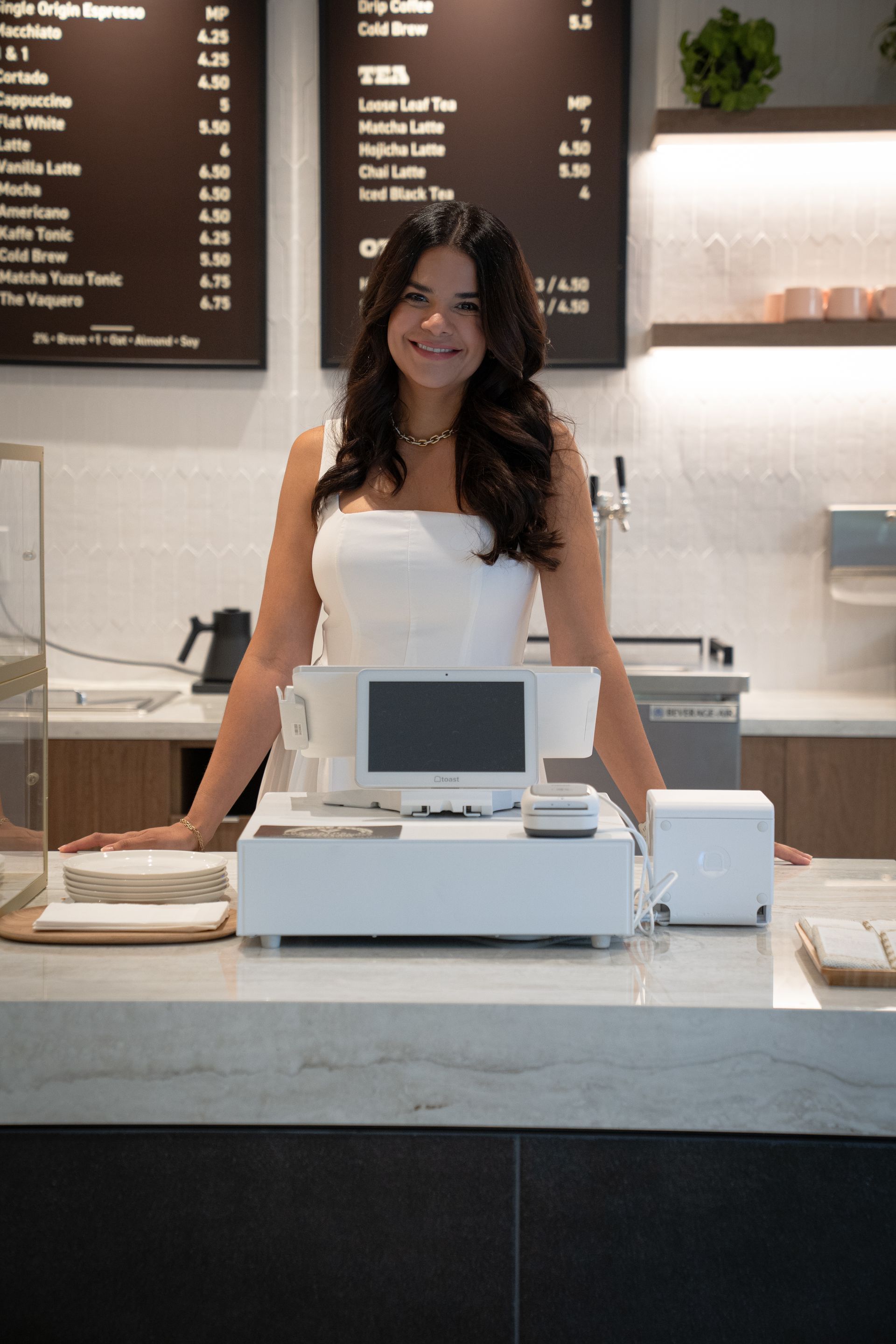 Barista smiling behind café counter with cash register and menu boards