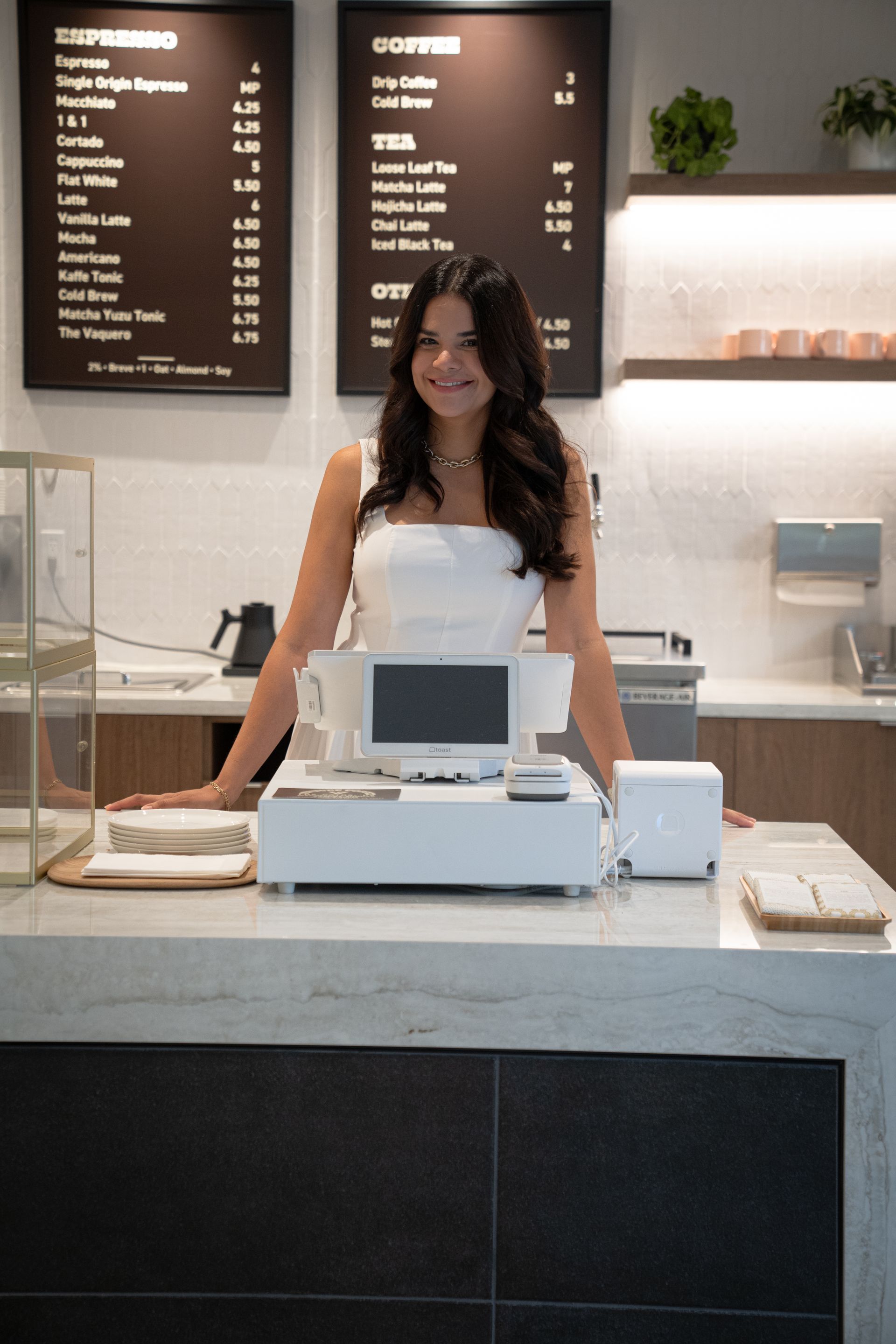 Smiling cashier behind a white counter in a modern cafe with menu boards in the background