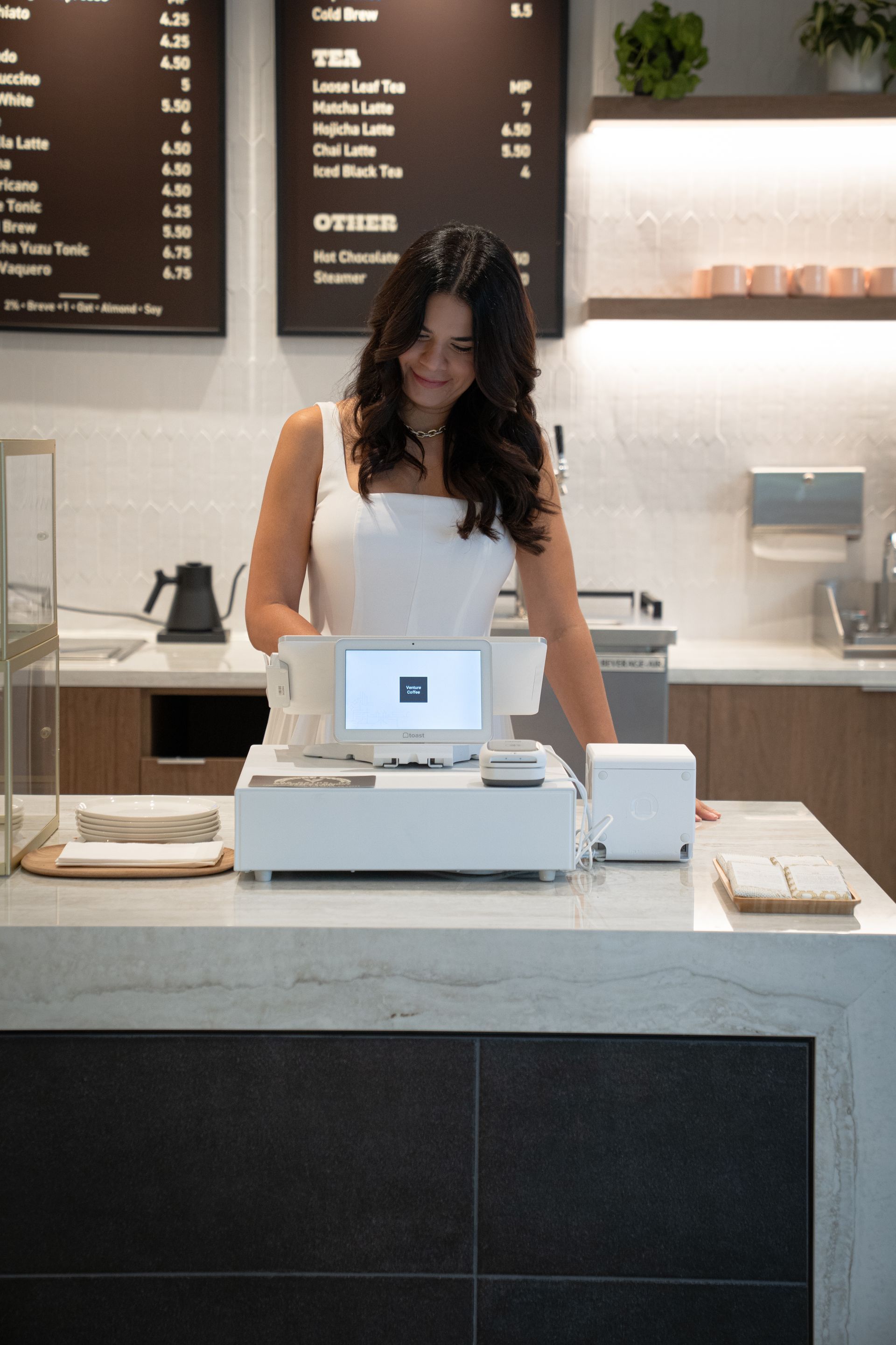 Barista serving at a café counter with a laptop register and menu board behind her