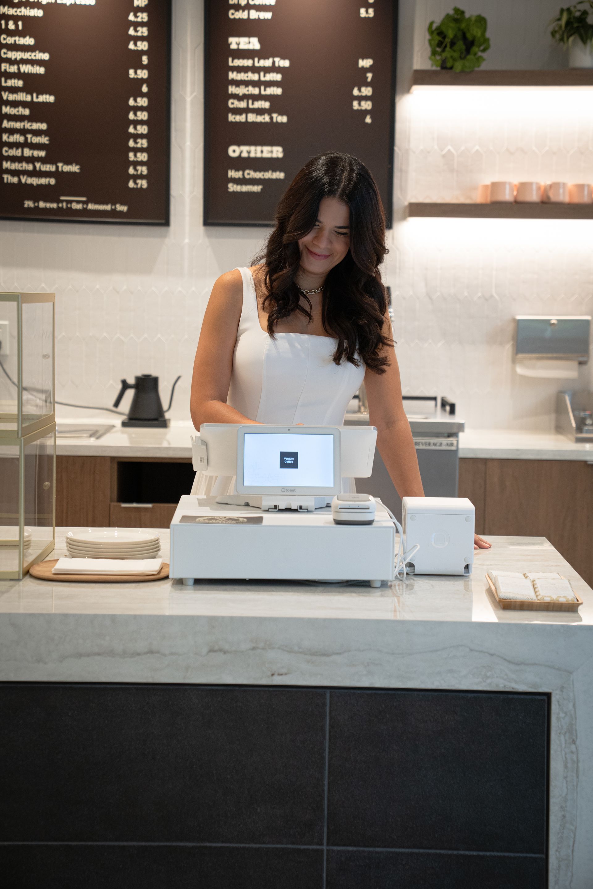 Barista behind café counter preparing an order with a white register and menu boards in the background