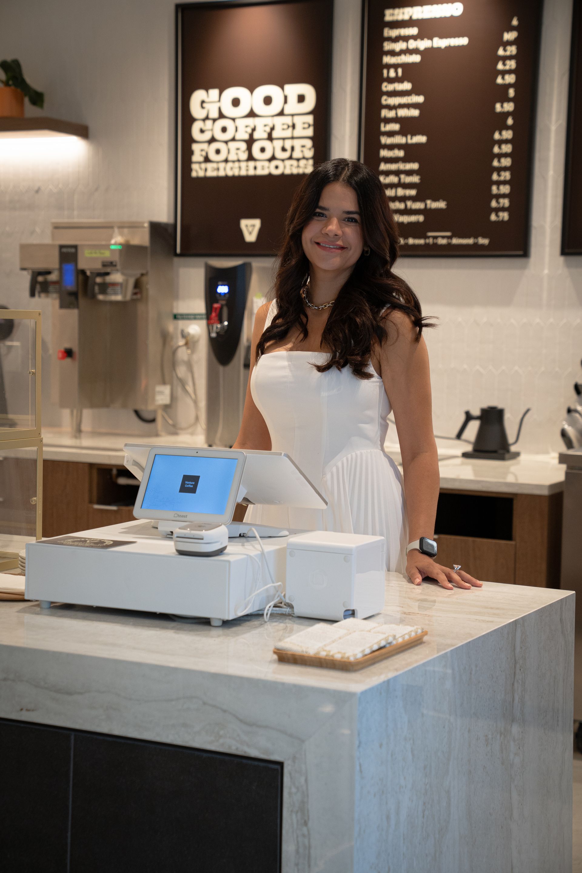Barista smiling behind a café counter with coffee equipment and menu boards in the background