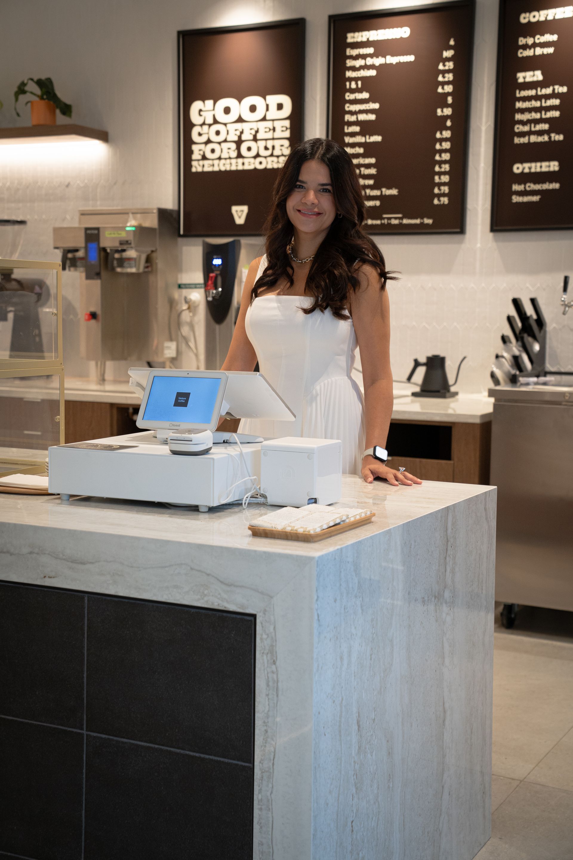 Woman standing behind a café counter with a point-of-sale tablet and menu boards in the background