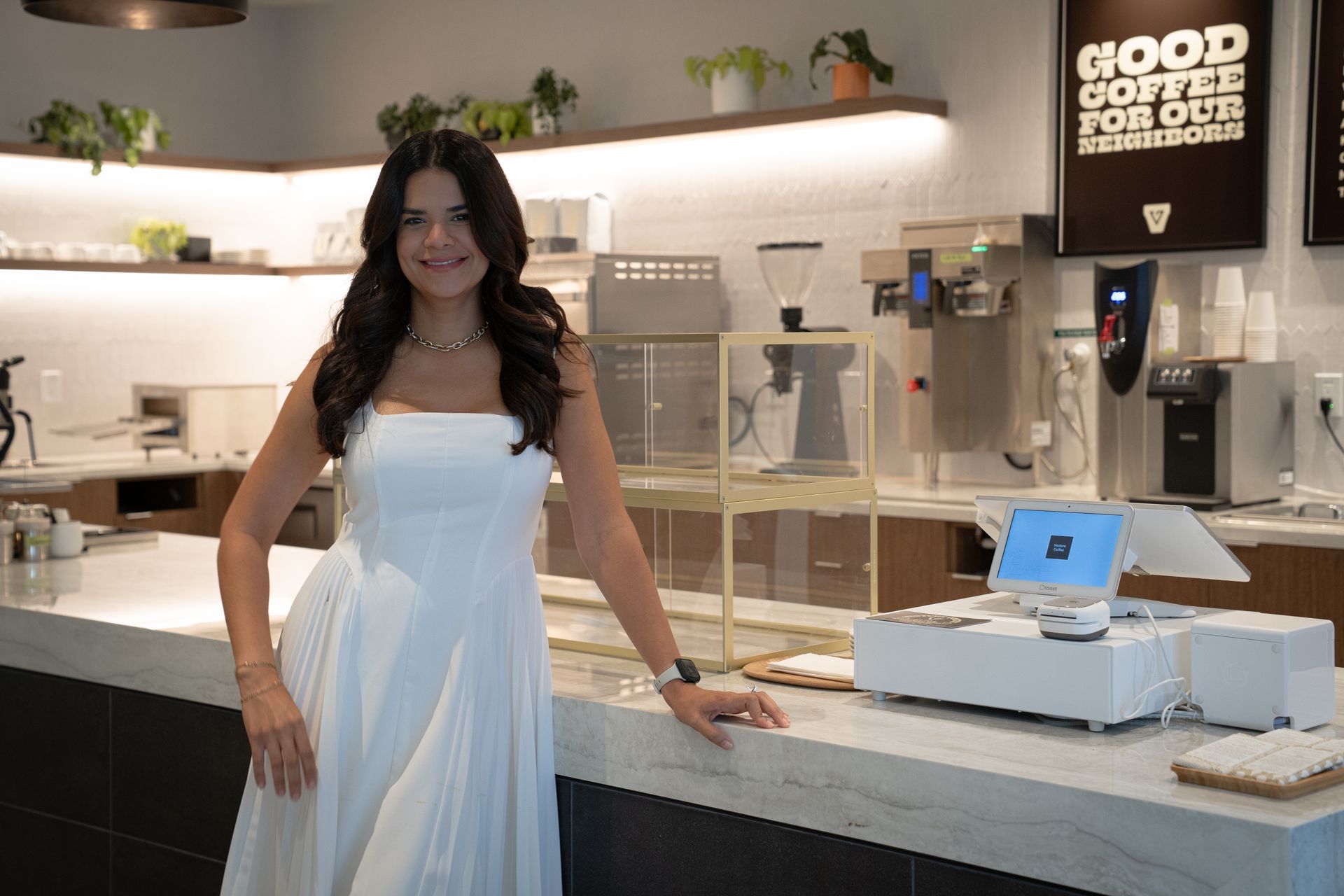 Woman in a white dress standing at a cafe counter with coffee machines and a 