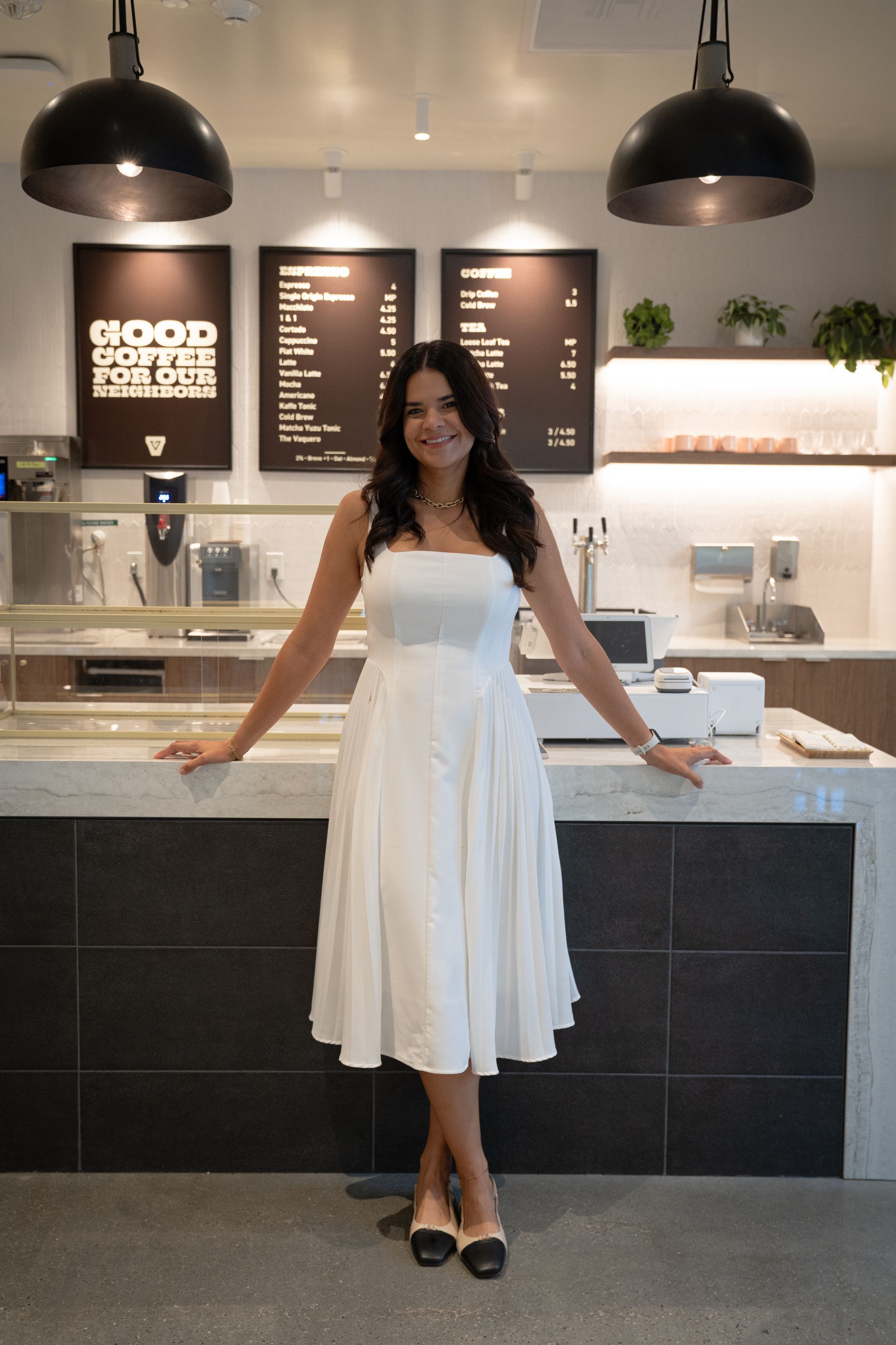 Woman in a white dress standing at a cafe counter with pendant lights and menu boards behind her