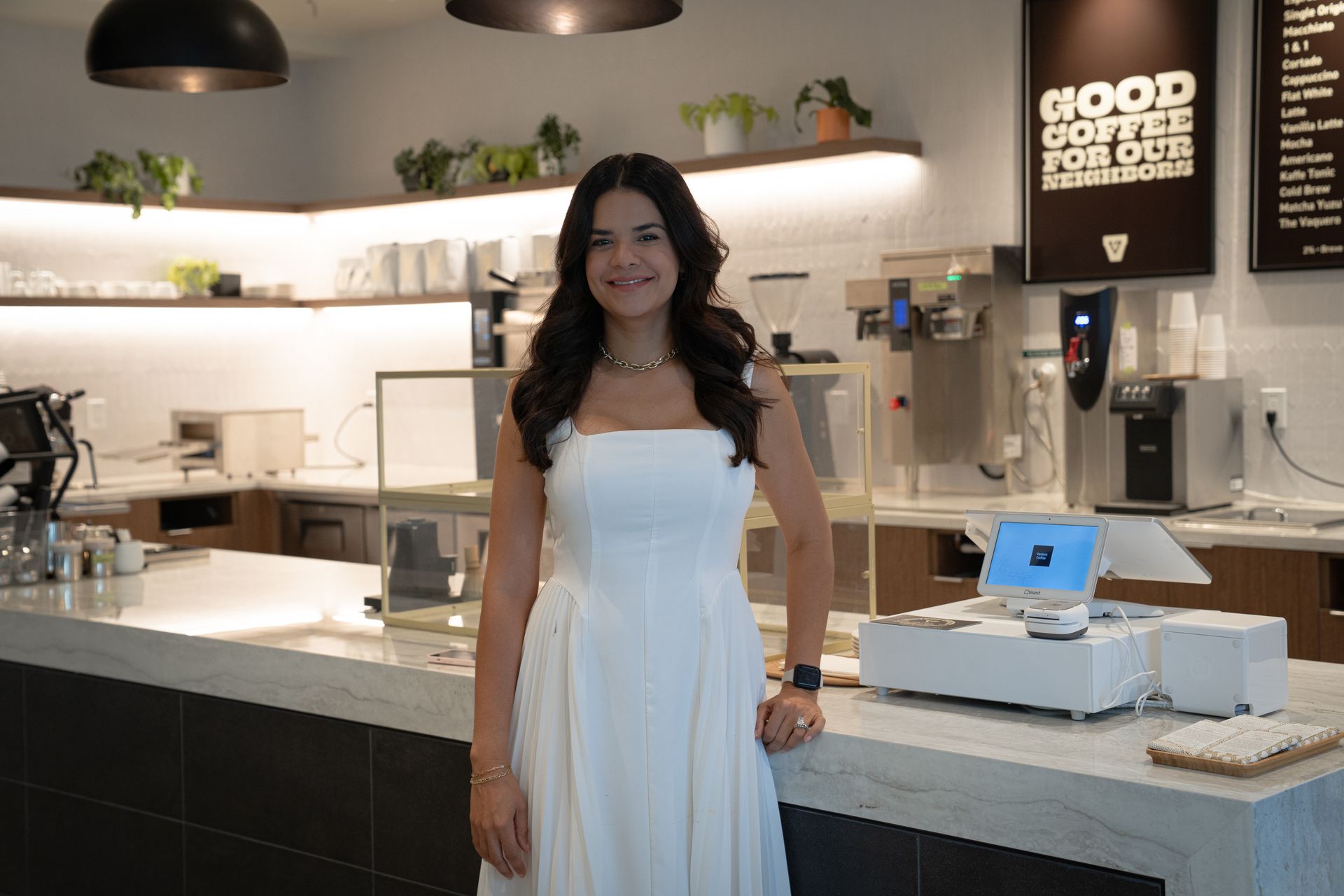 Woman in a white dress standing behind a counter in a modern café or dessert shop.