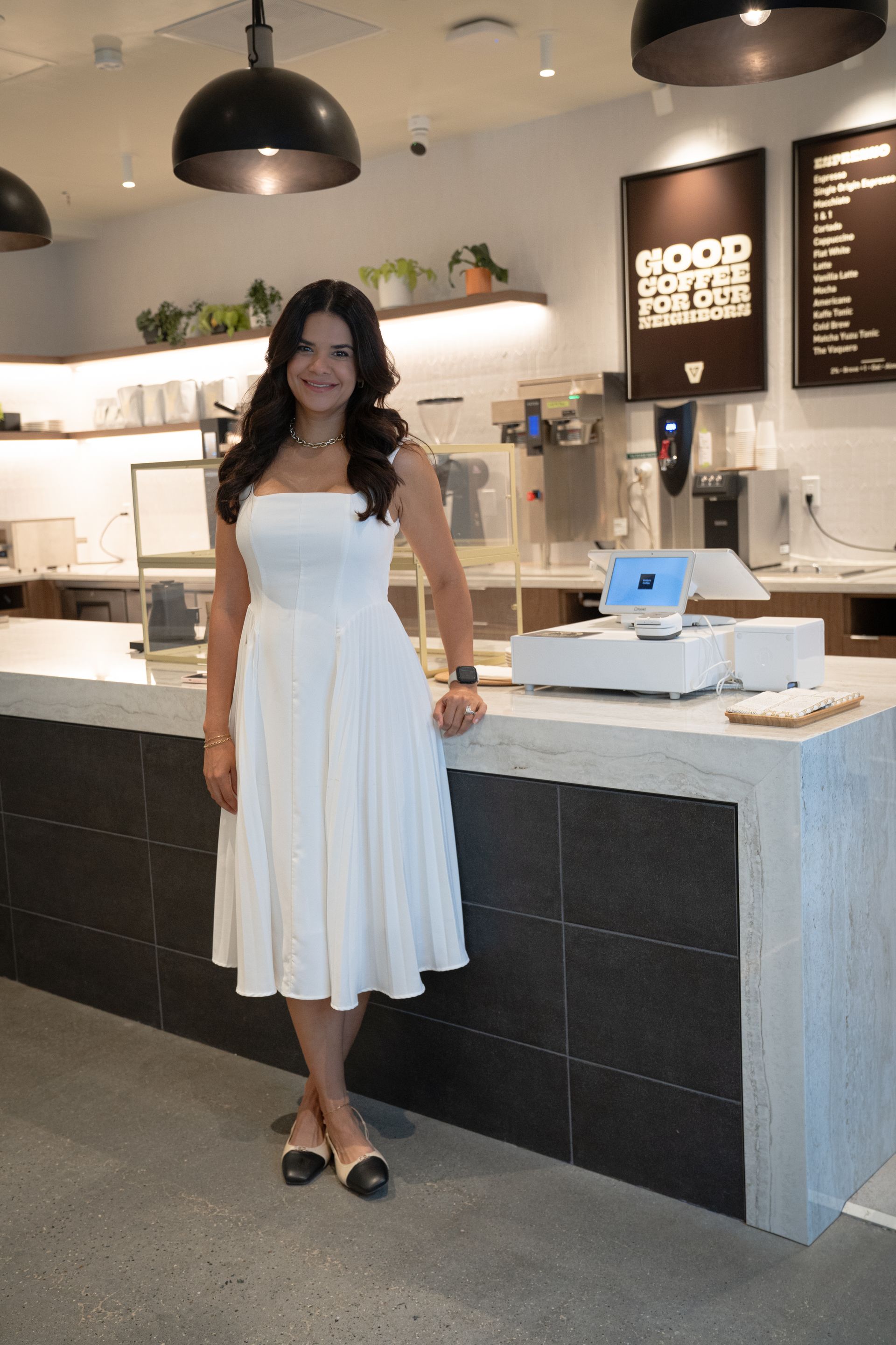 Woman in a white dress standing in a café beside a counter and register.
