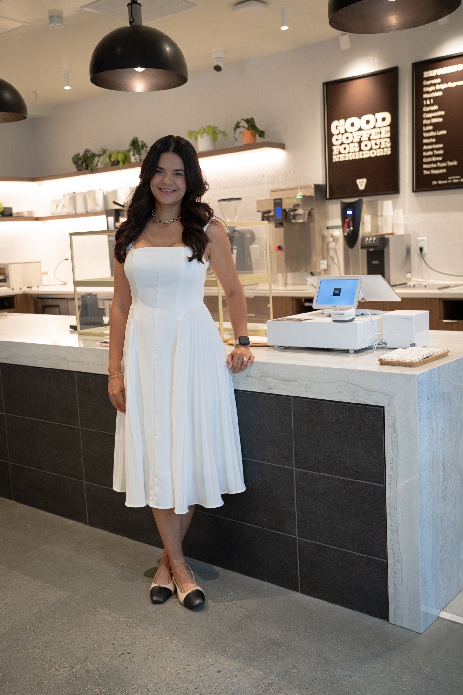 Woman in a white dress standing in a café with a counter and hanging lights behind her