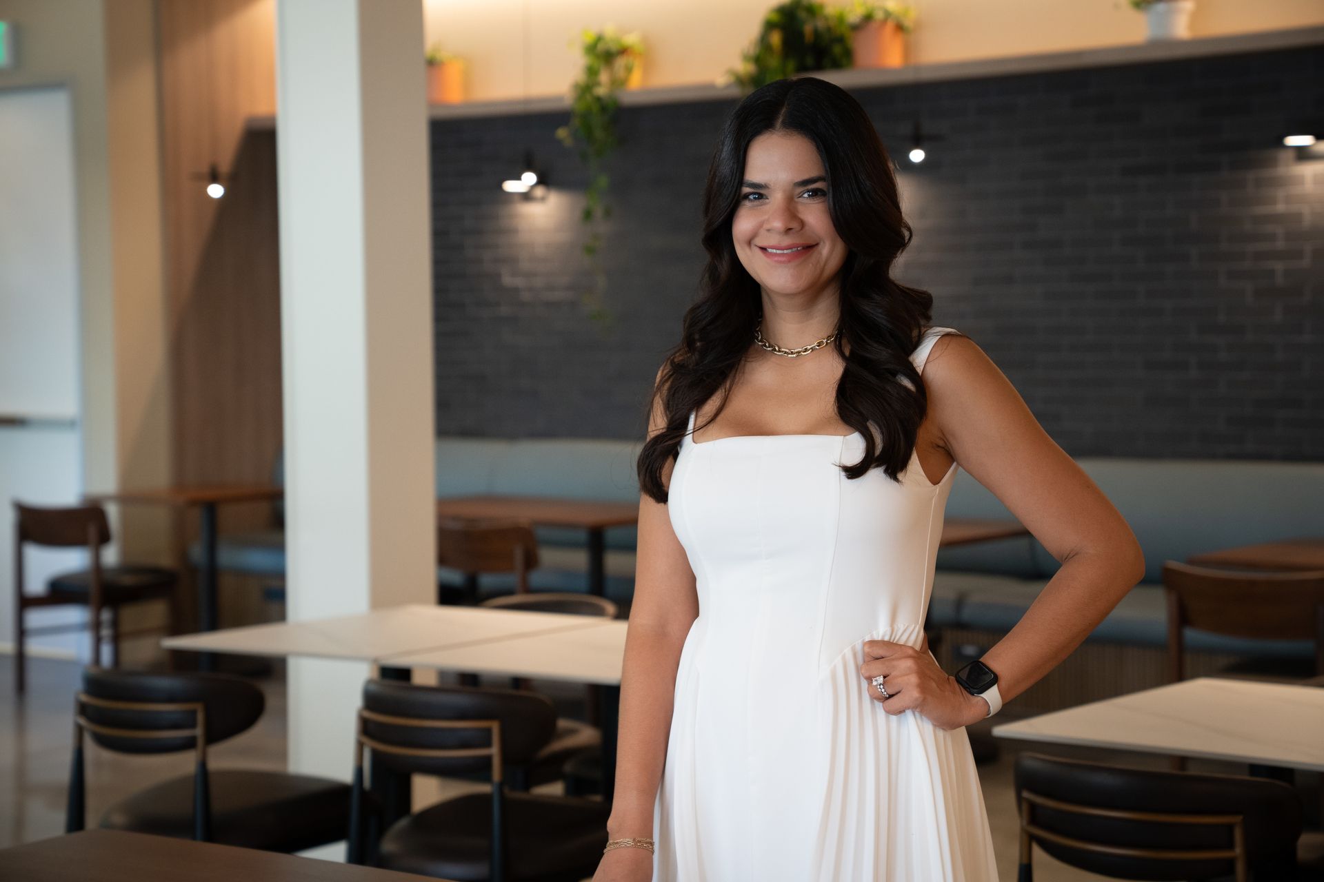 Woman in a white dress standing in a modern café, smiling with hand on hip.