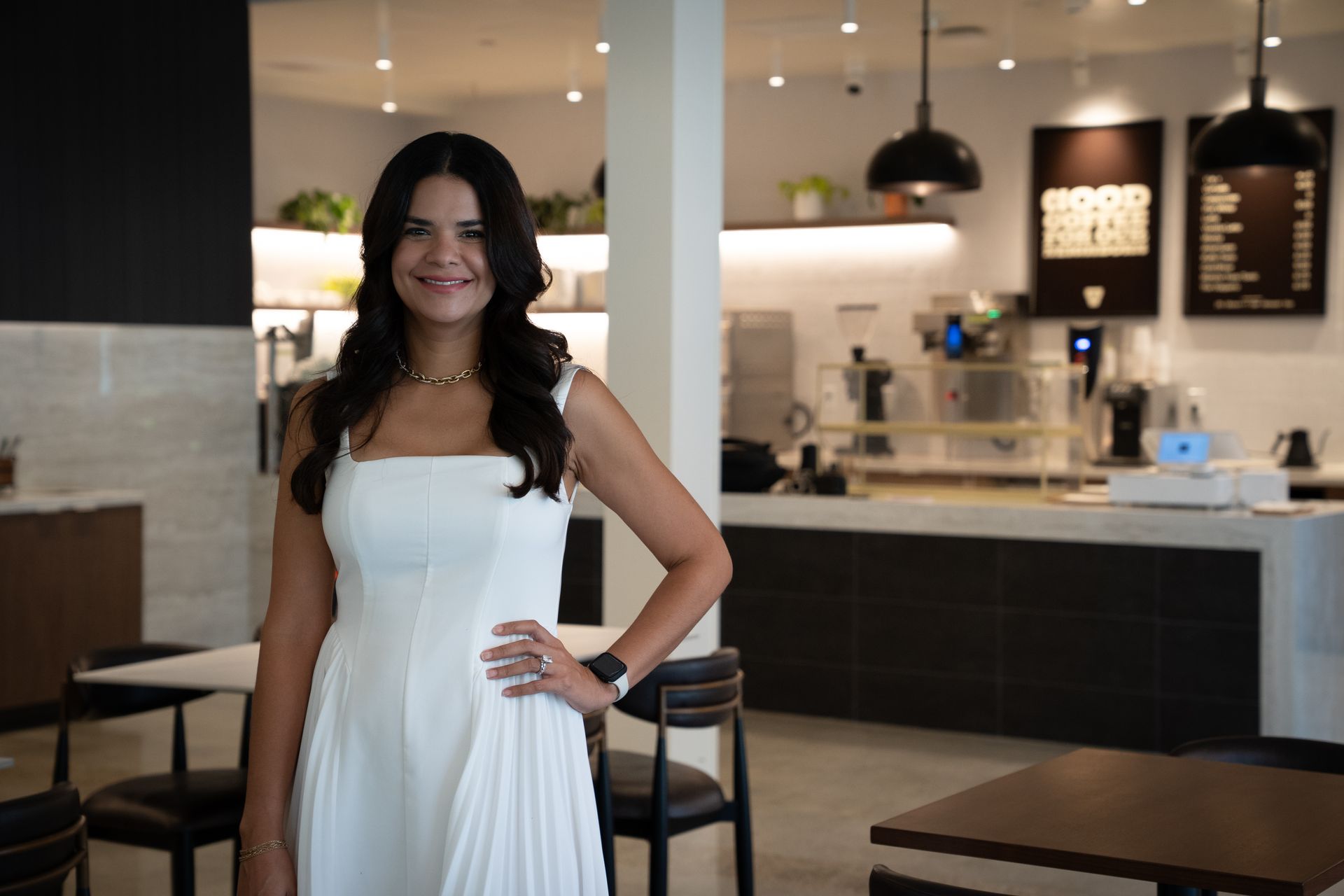 Woman in a white dress standing in a modern cafe with hand on hip, smiling.