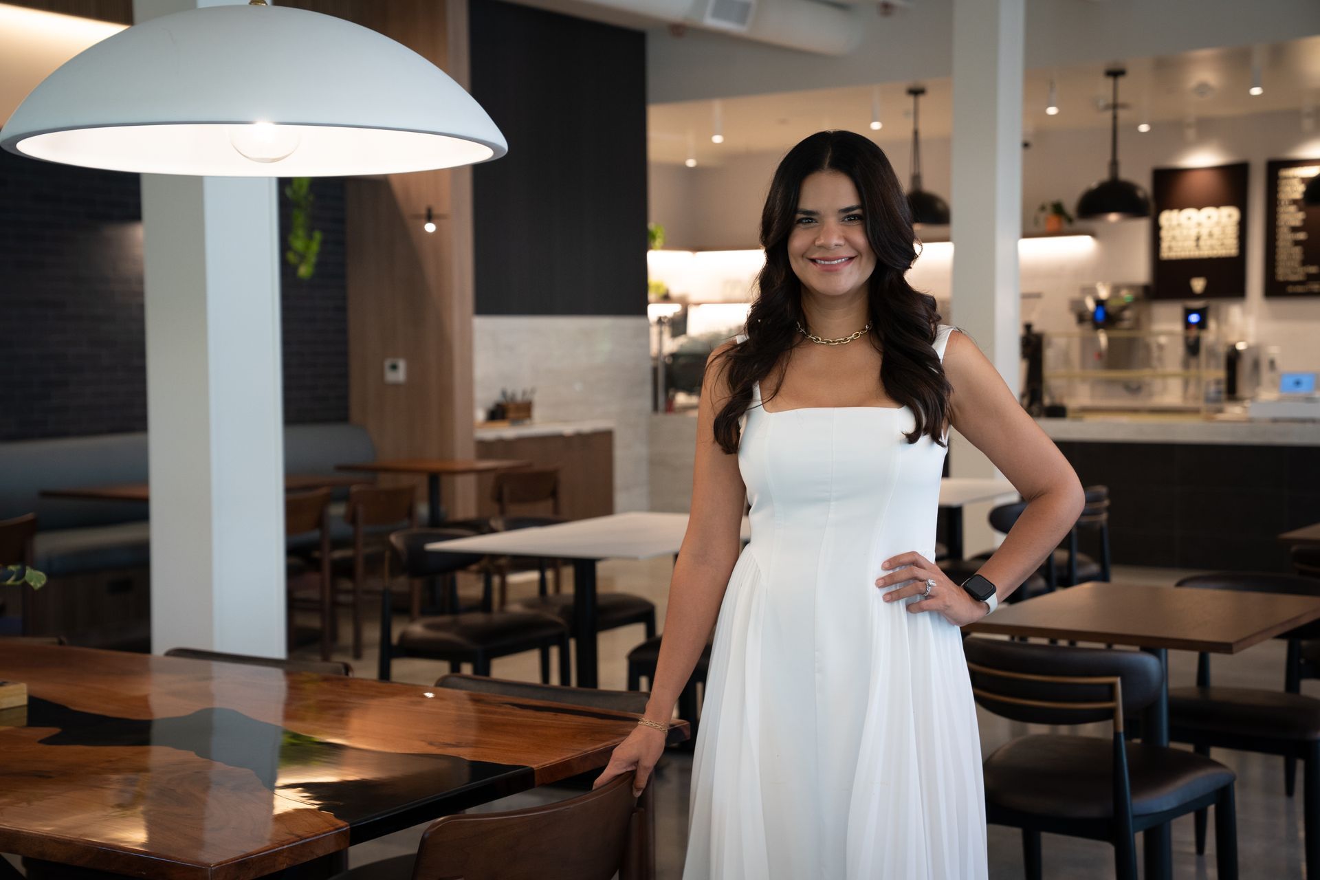 Woman in a white dress standing in a modern restaurant, smiling with one hand on her hip.