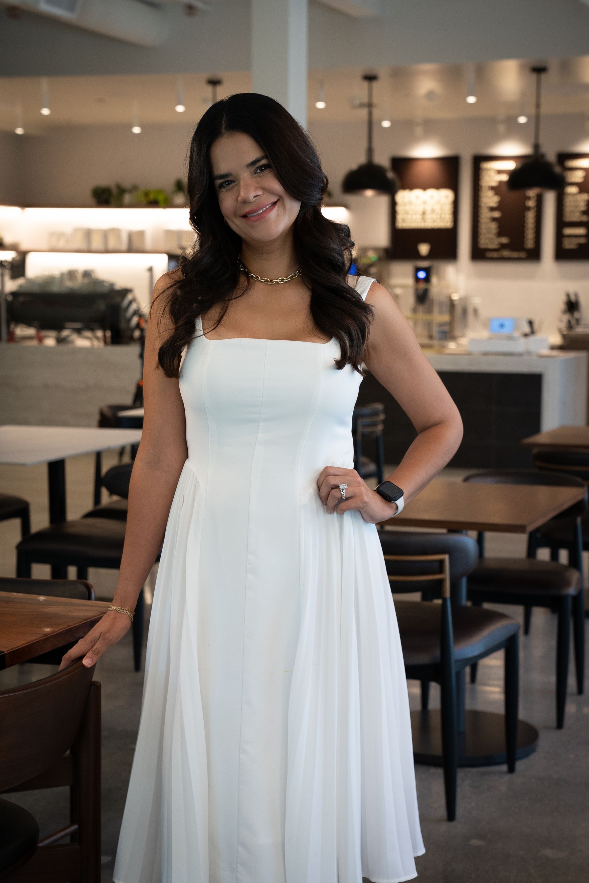 Woman in a white dress posing indoors at a restaurant, smiling beside wooden tables.