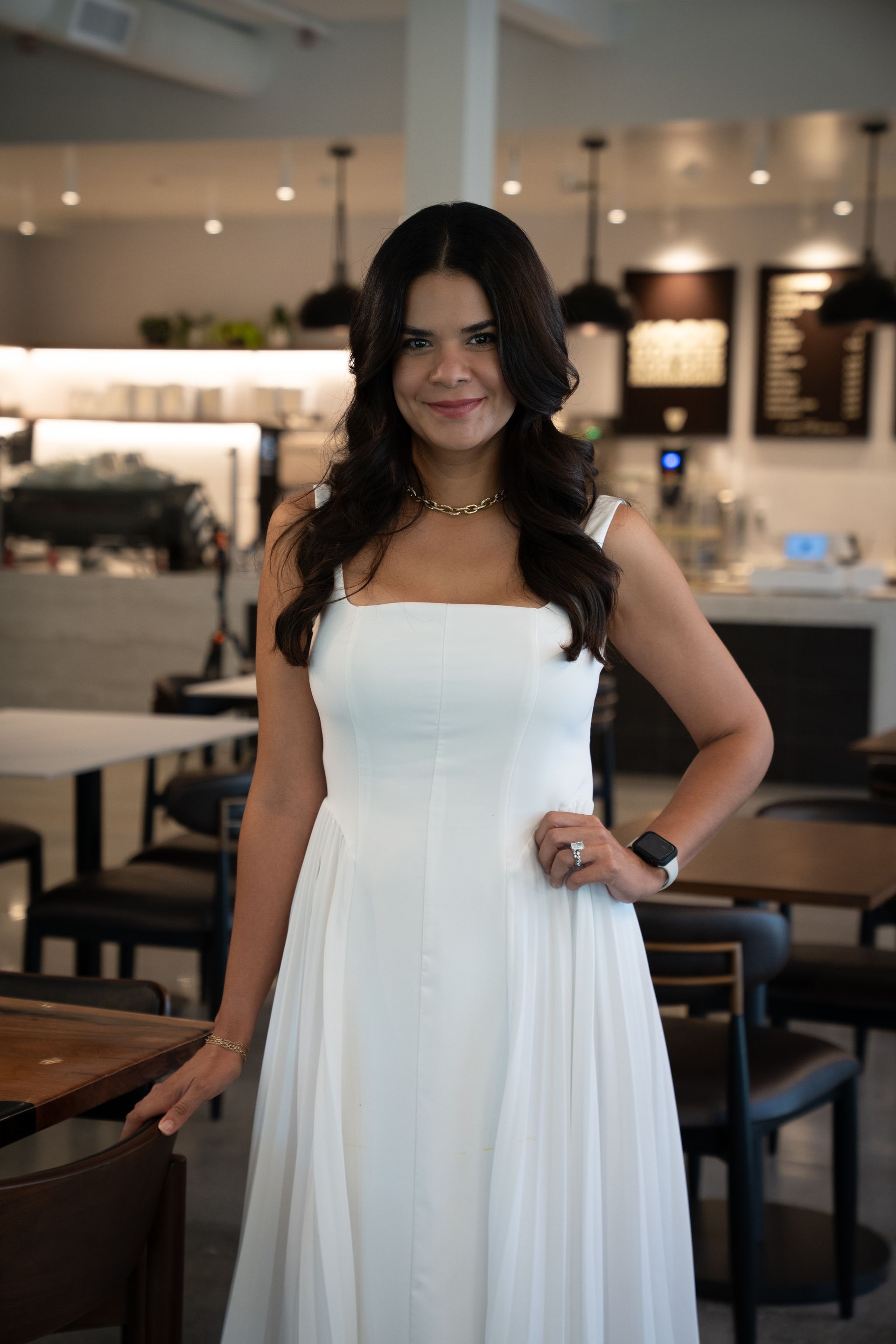 Woman in a white dress posing in a modern café, standing with one hand on her hip.