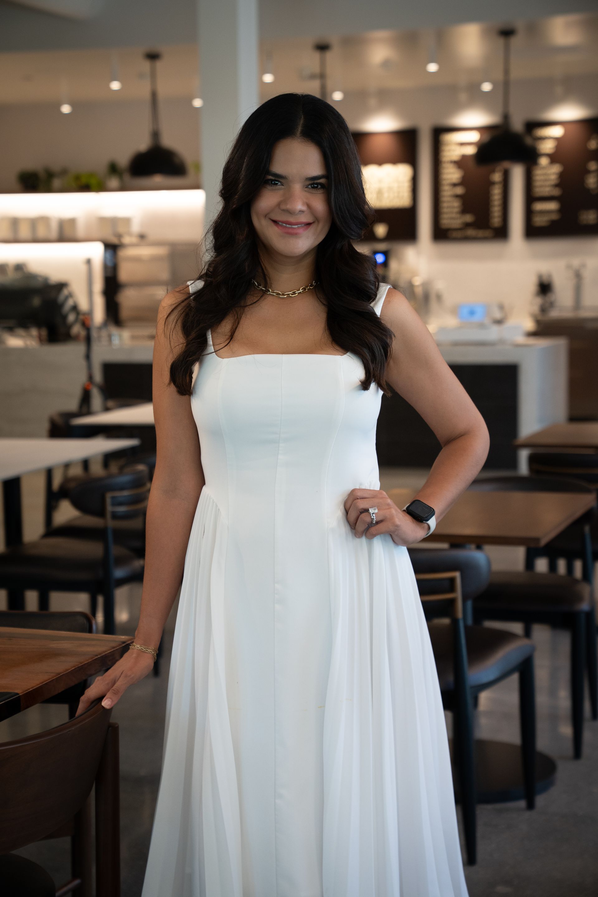 Woman in a white dress standing in a softly lit restaurant, smiling with one hand on her hip.