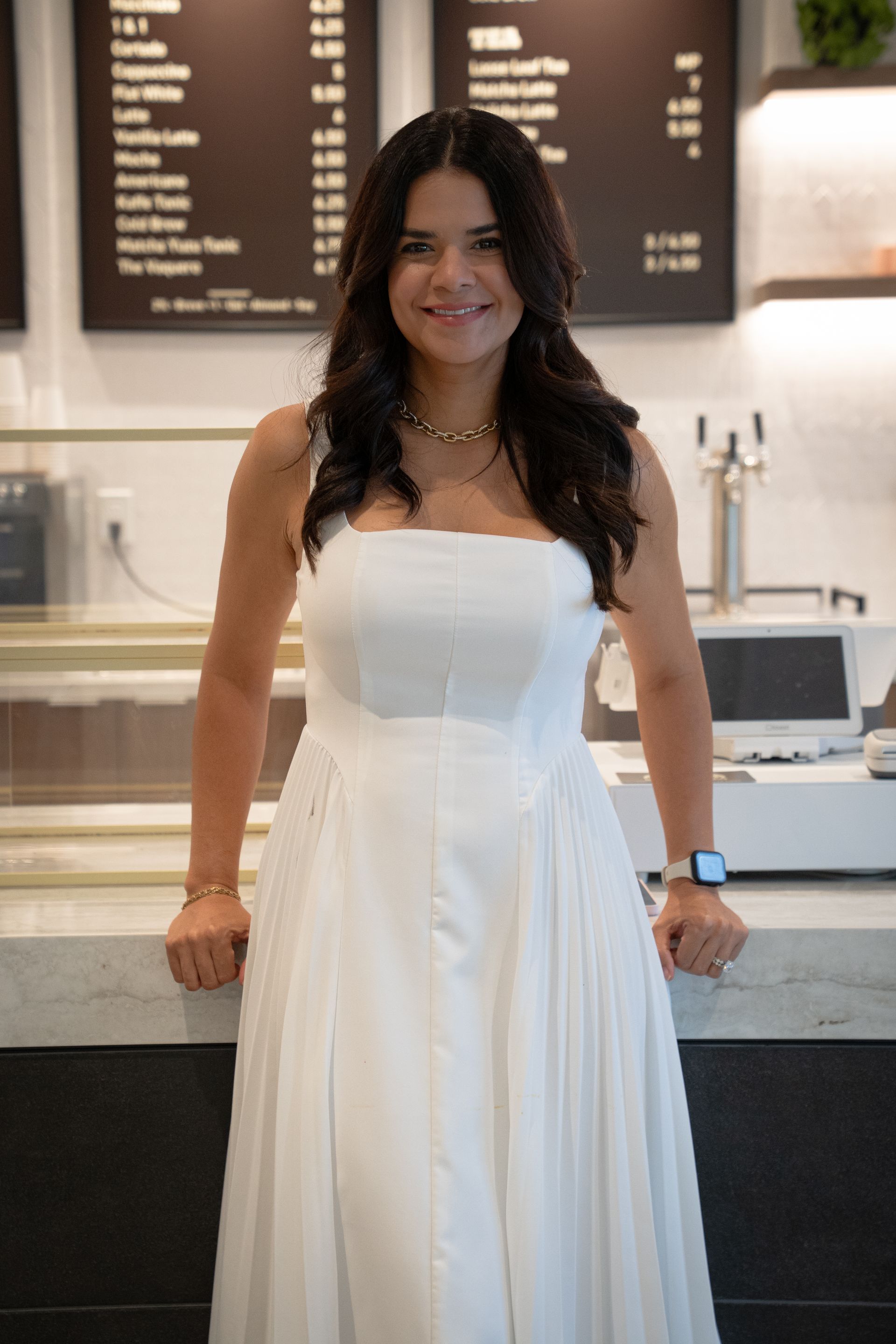 Woman in a white dress standing in a café, smiling at the counter.