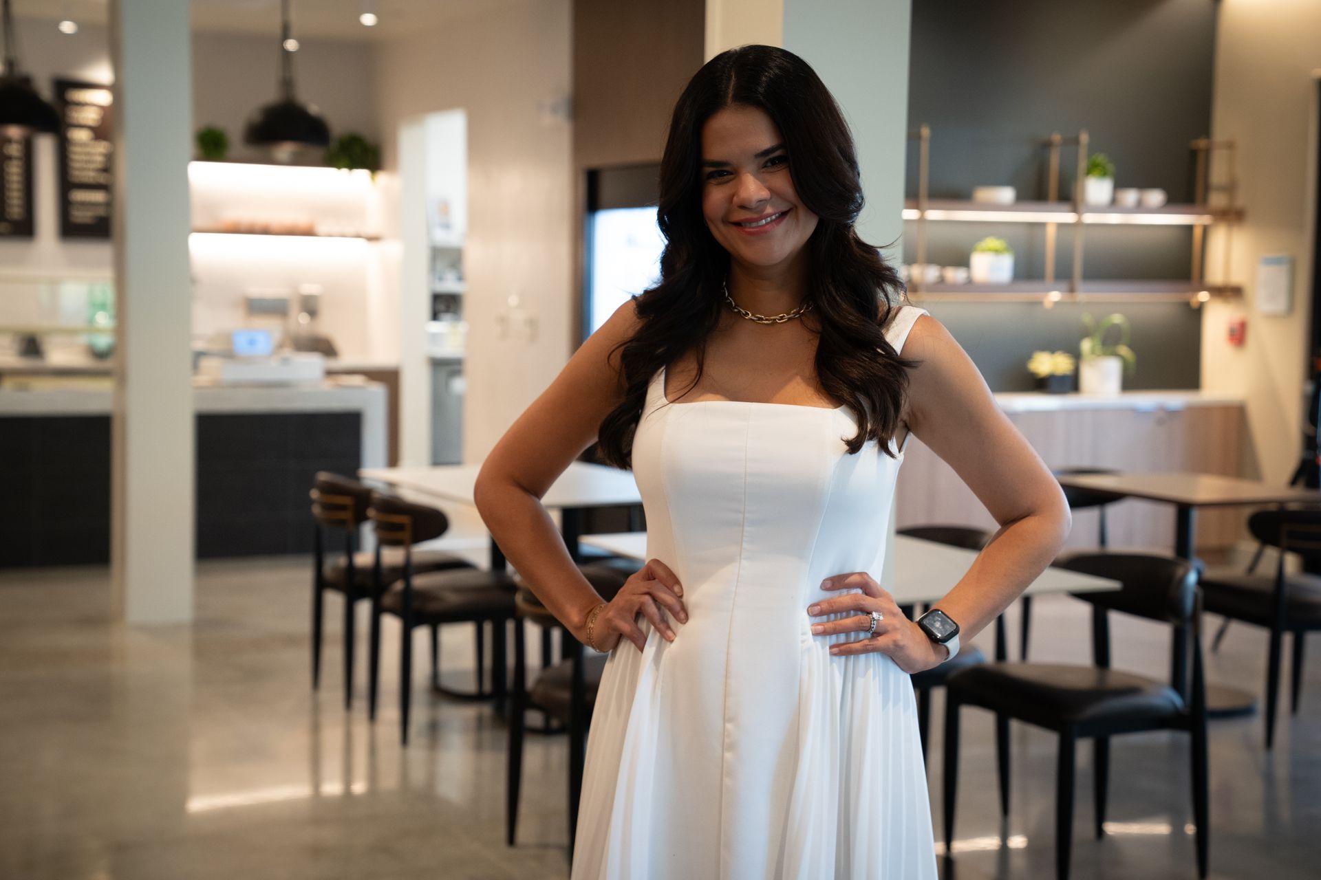 Woman in a white dress standing in a modern kitchen, hands on hips and smiling.