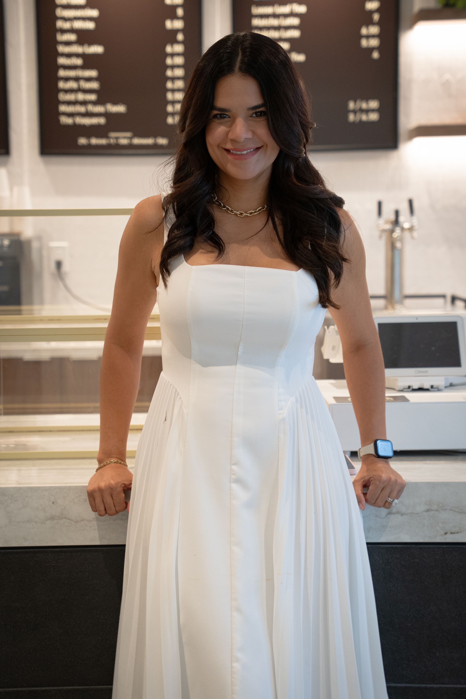 Woman in a white dress smiling at a café counter, with menu boards in the background.