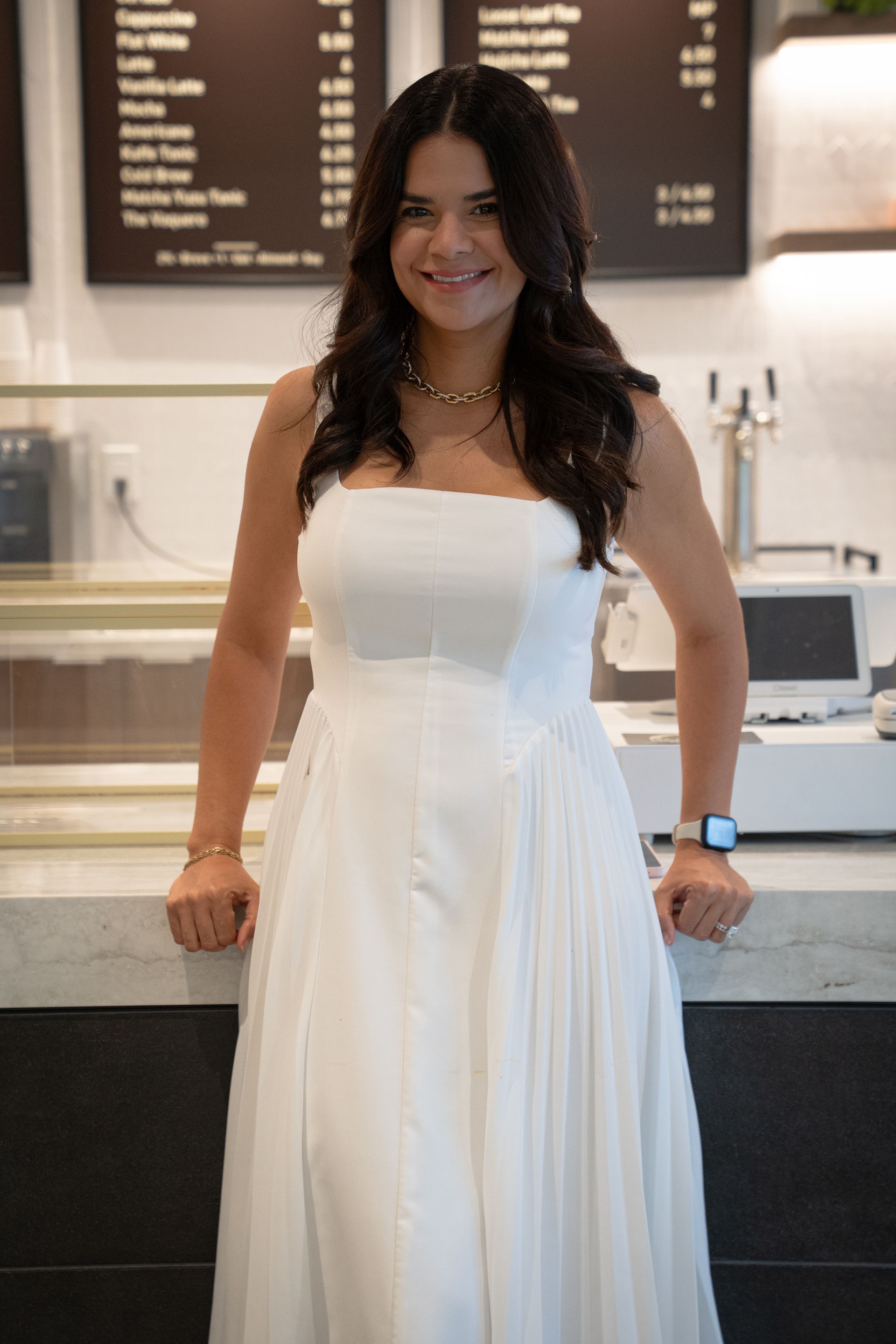 Woman in a white strapless dress standing at a cafe counter, smiling with hands on the counter.