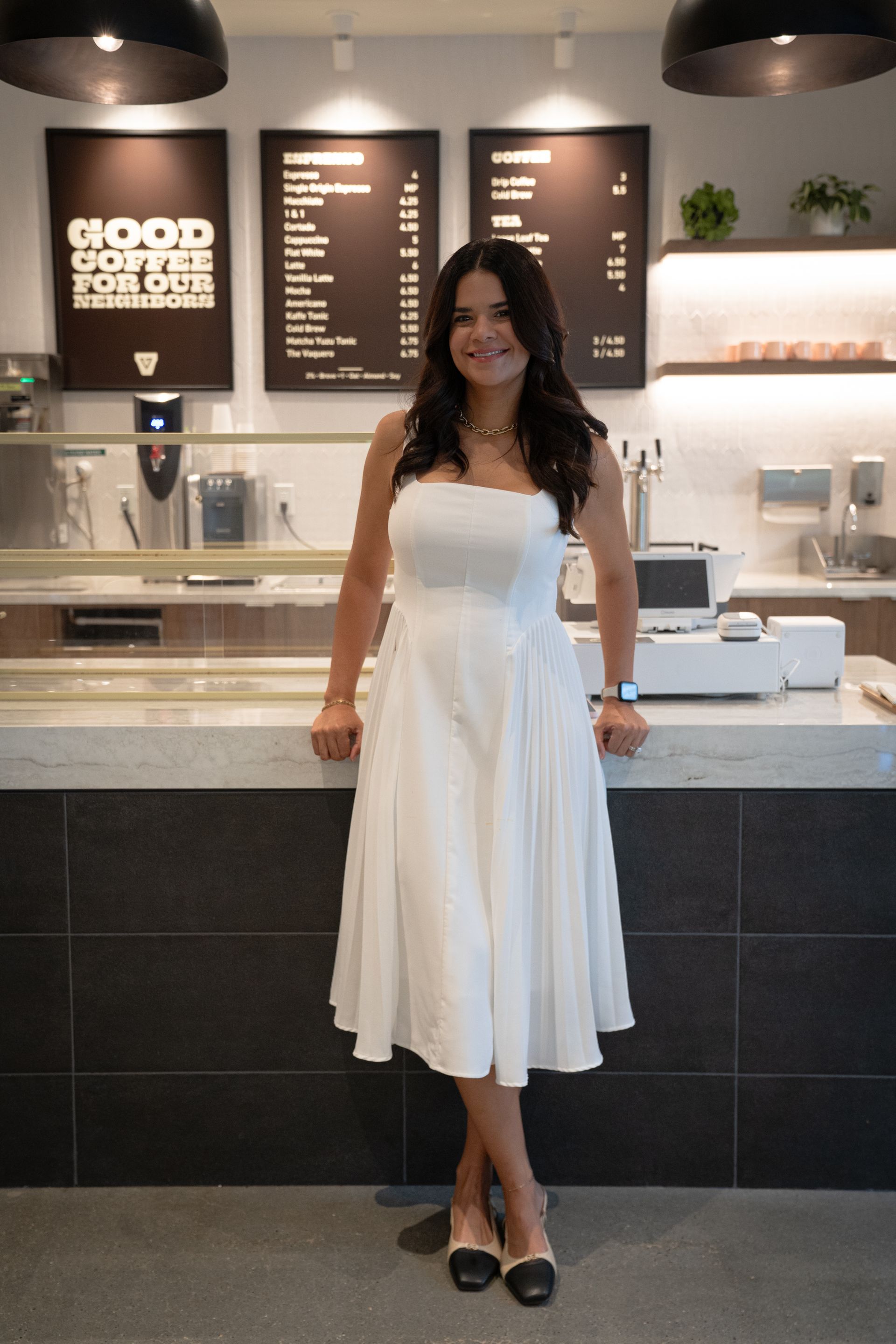 Woman in a white dress standing in a café with a menu board and counter behind her