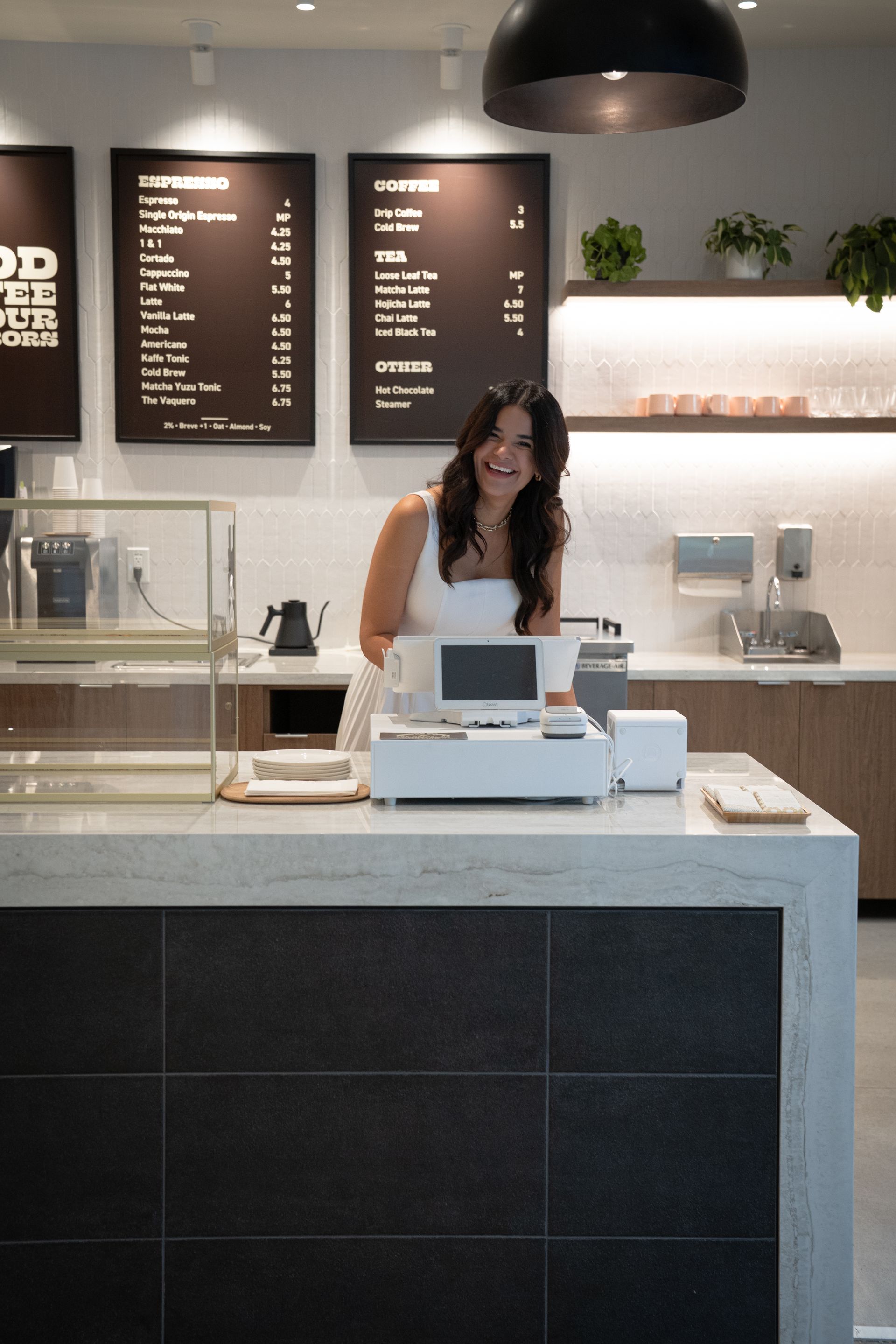 Barista at a modern café counter with menu boards, plants, and a coffee machine خلف her
