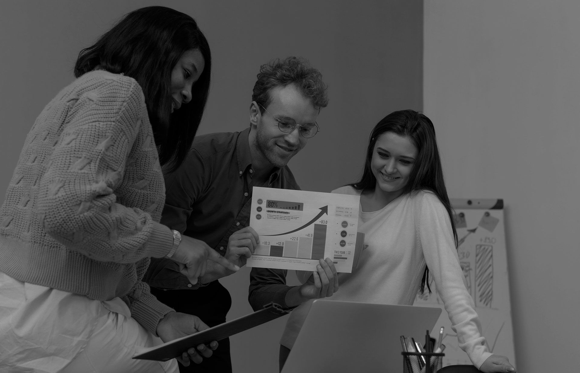 Three coworkers review a colorful chart together at a laptop in an office.