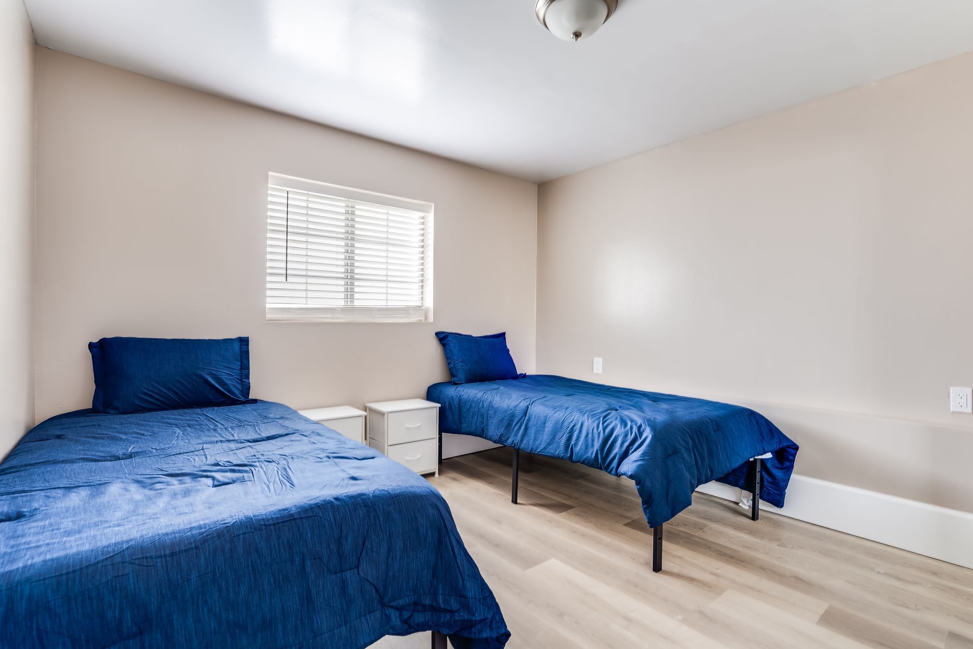 Two twin beds with blue bedding in a bedroom with beige walls and a window.