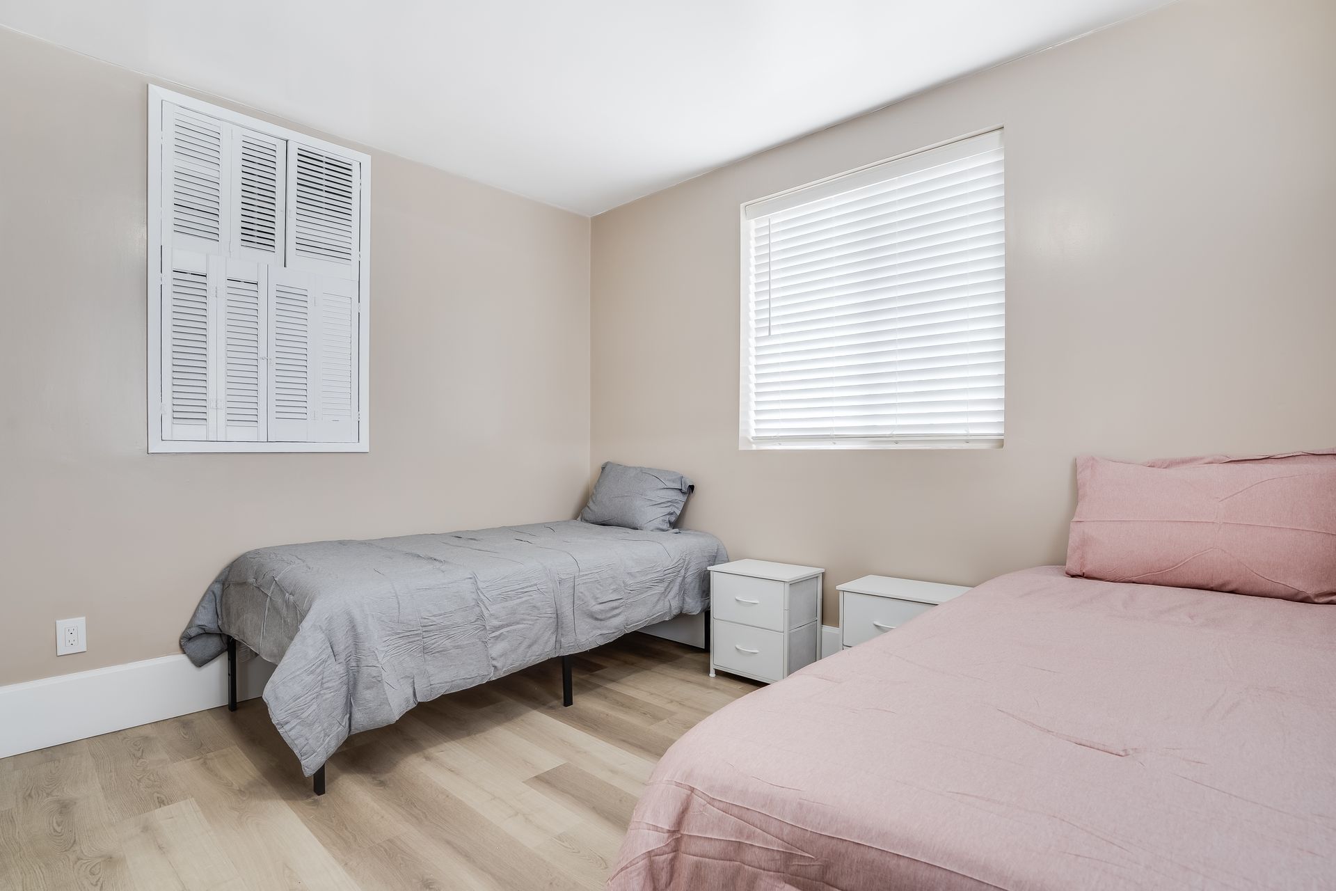 Two twin beds in a bedroom. One has gray bedding, the other pink. Neutral walls, artwork, and blinds.