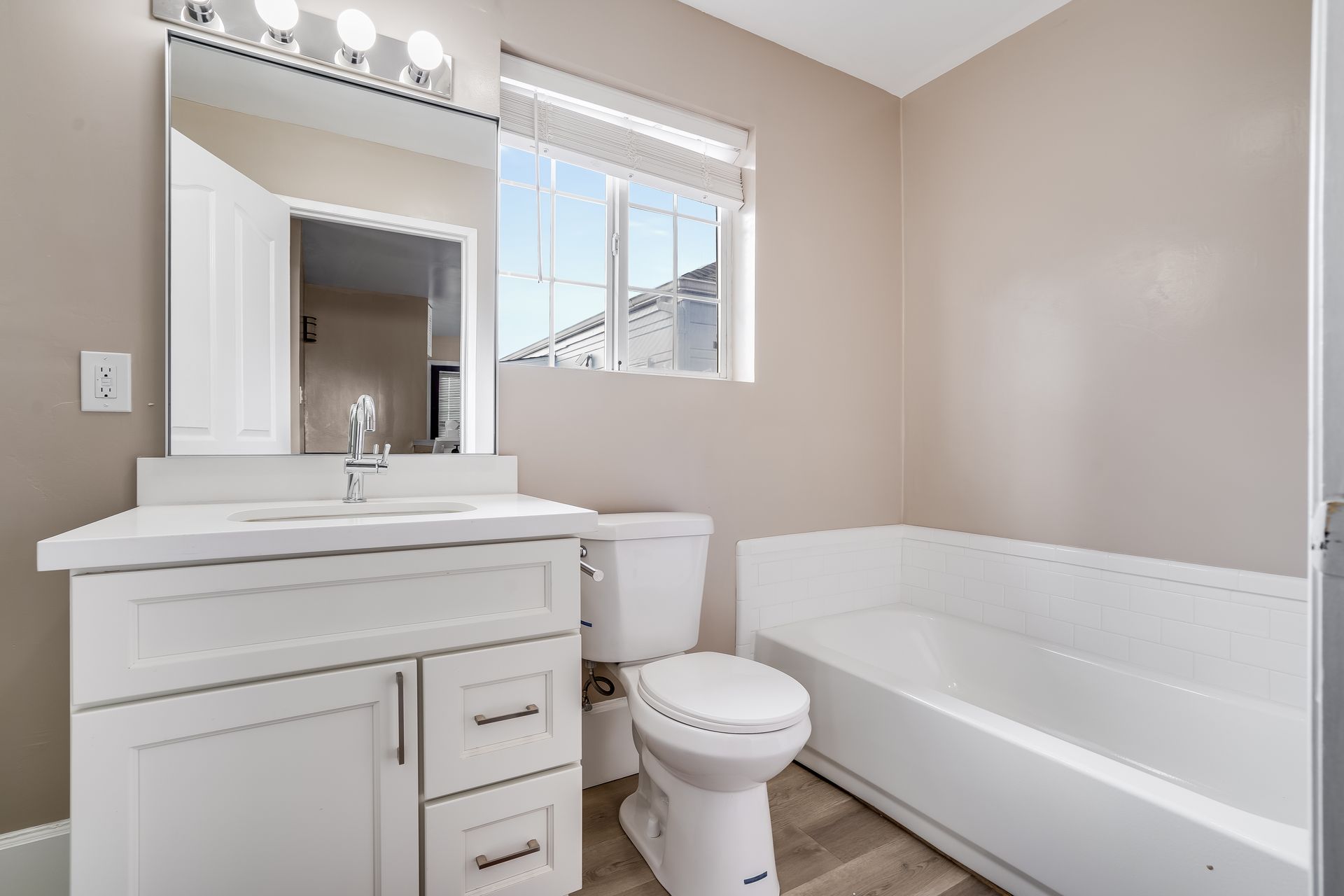 Bathroom with white vanity, toilet, and tub; tan walls, window with blinds.