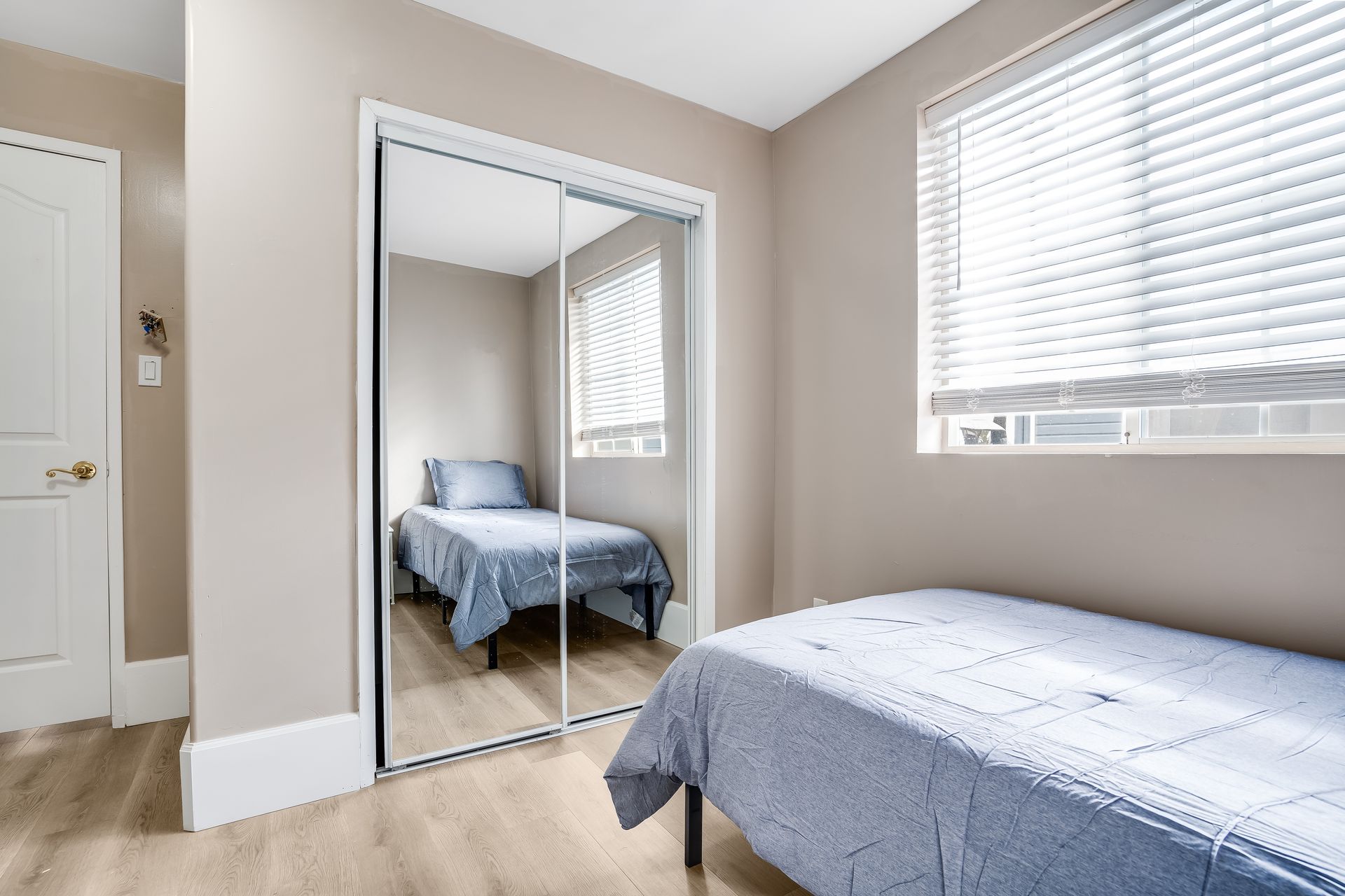 Bedroom with two twin beds, one reflected in a mirrored closet door. Light walls, wood floor, and blinds.