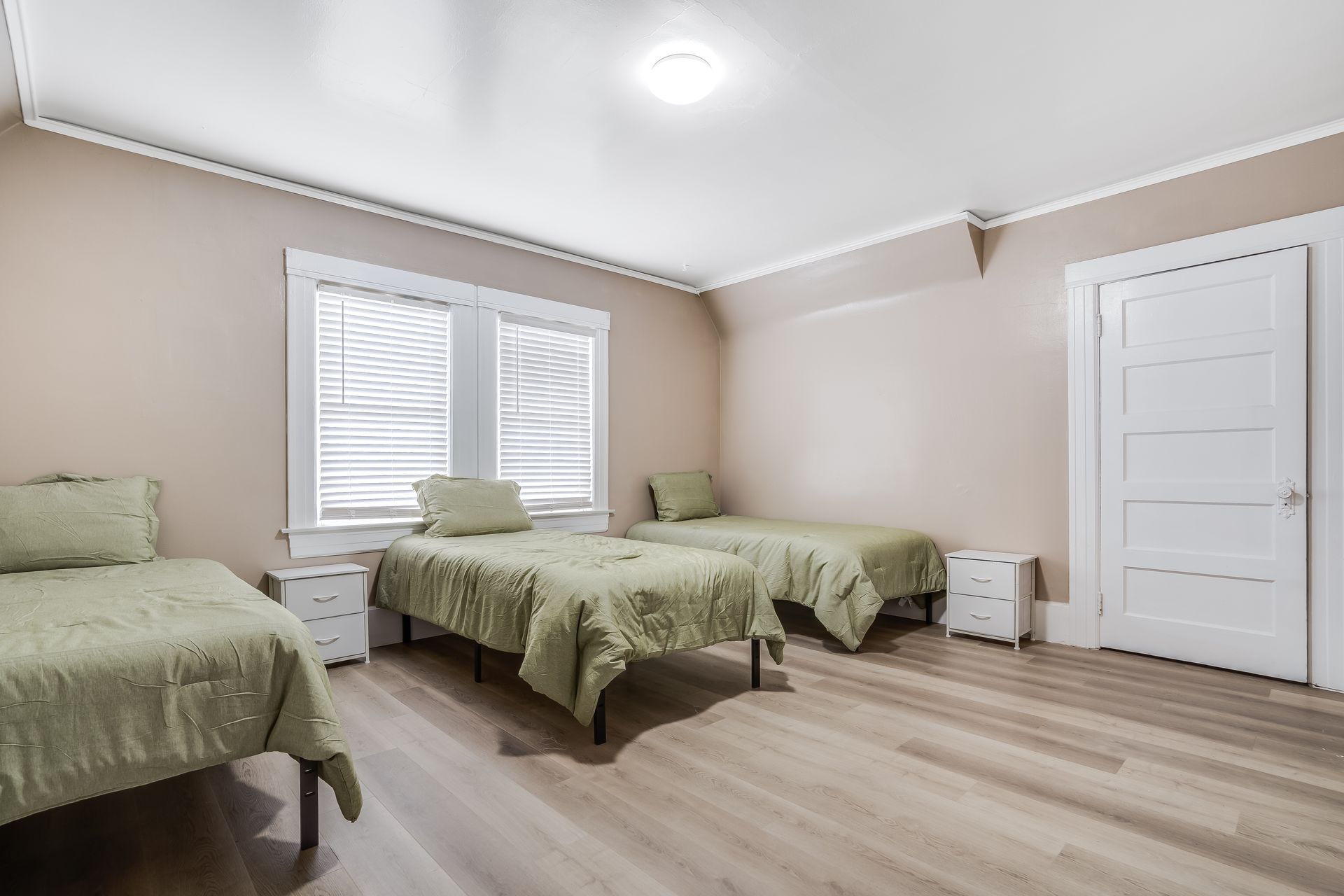 A bedroom with three beds, nightstands, a window, and a door. The room is painted beige, with light green bedding.