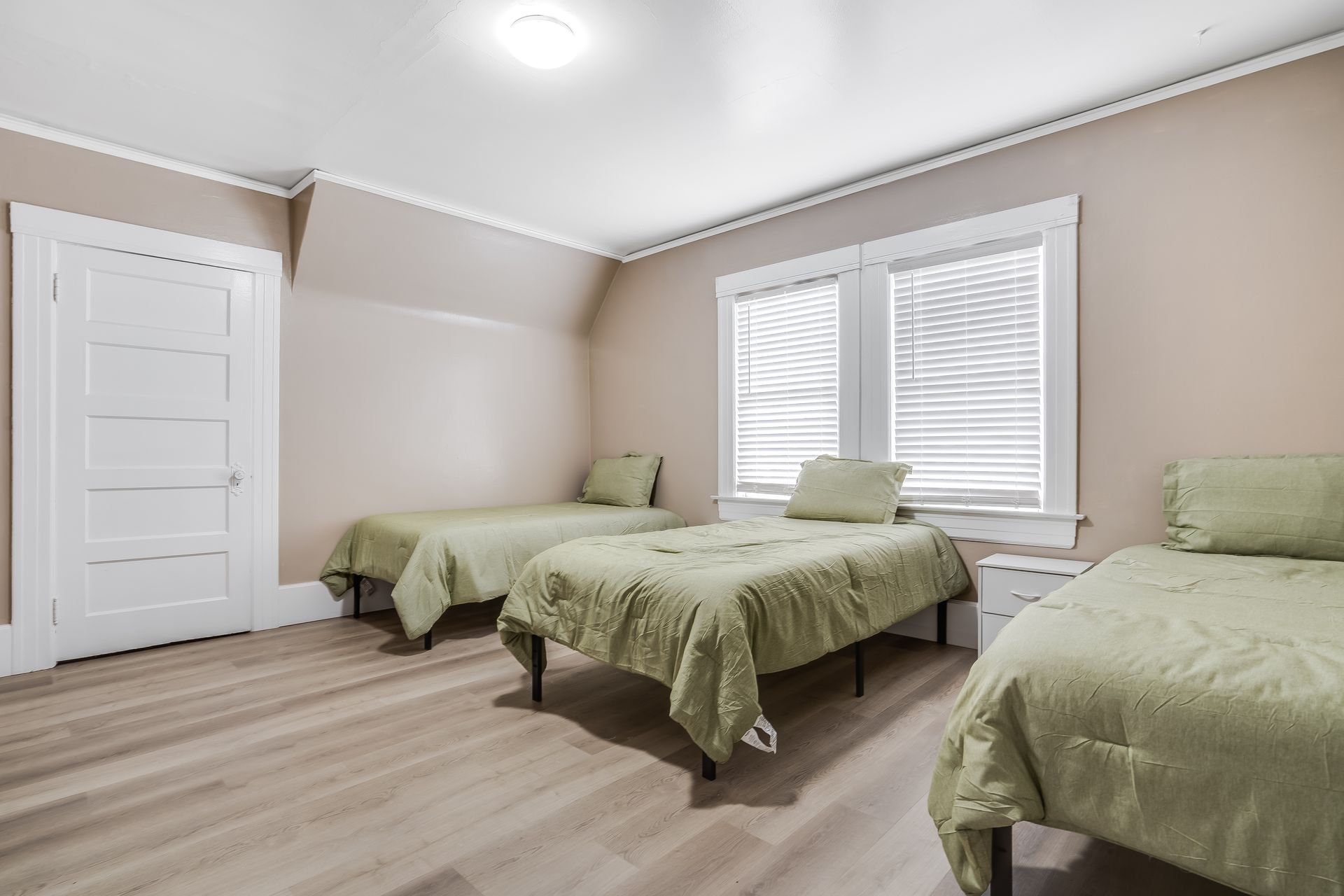 A bedroom with three beds, green bedding, and a white door and window. Light wood floors and tan walls.