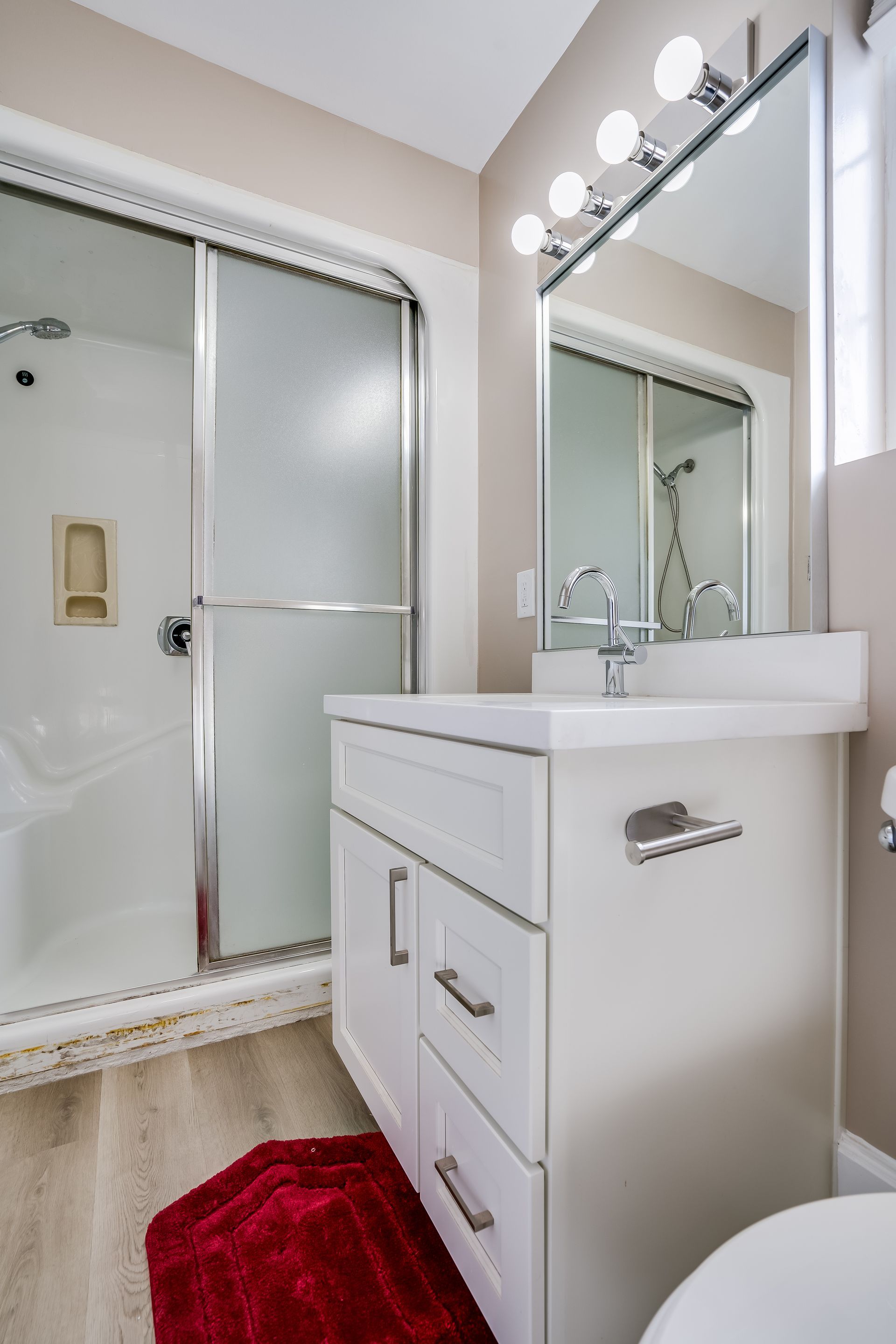 Bathroom with white vanity, shower with frosted glass door, red rug, and mirror.