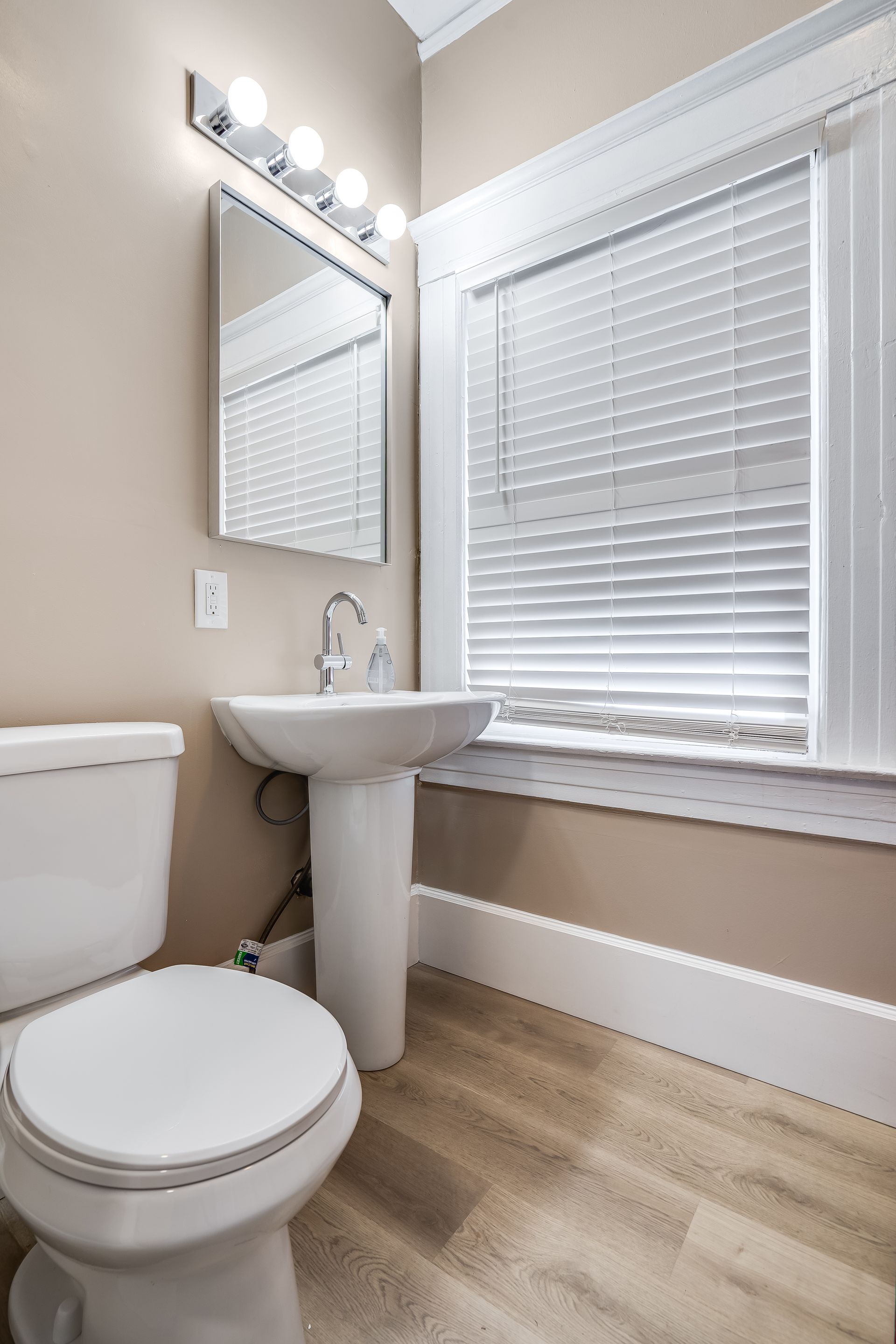 Bathroom with a toilet, pedestal sink, mirror, and window with blinds. Beige walls and light wood floor.