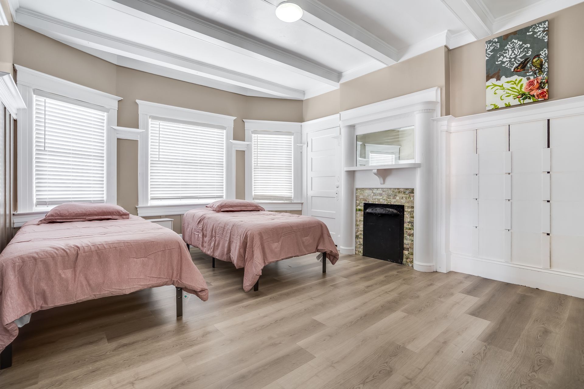 Bedroom with two beds, white trim, beige walls, faux fireplace, and wood flooring.
