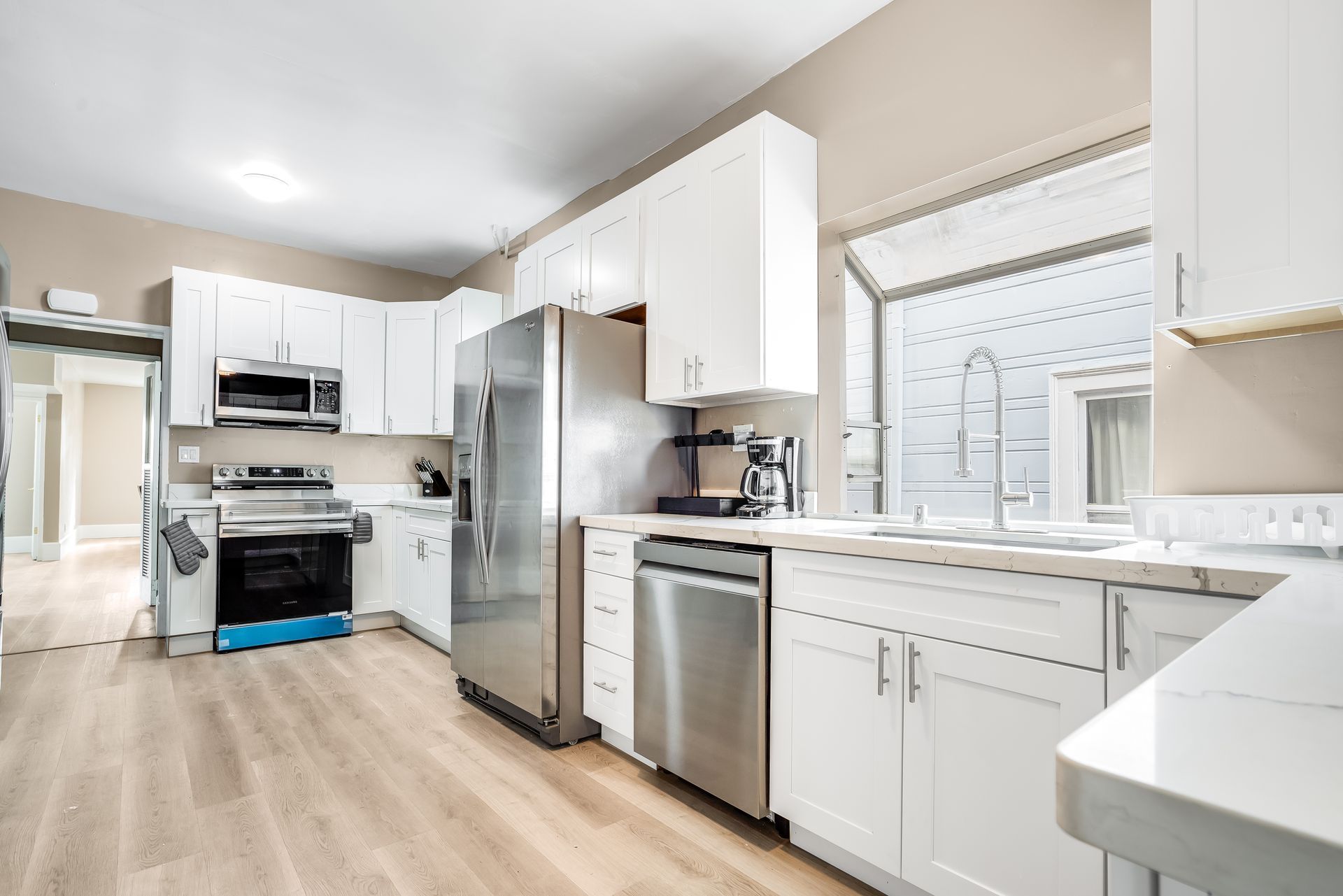 Bright white kitchen with stainless steel appliances, light wood floors, and a large window.