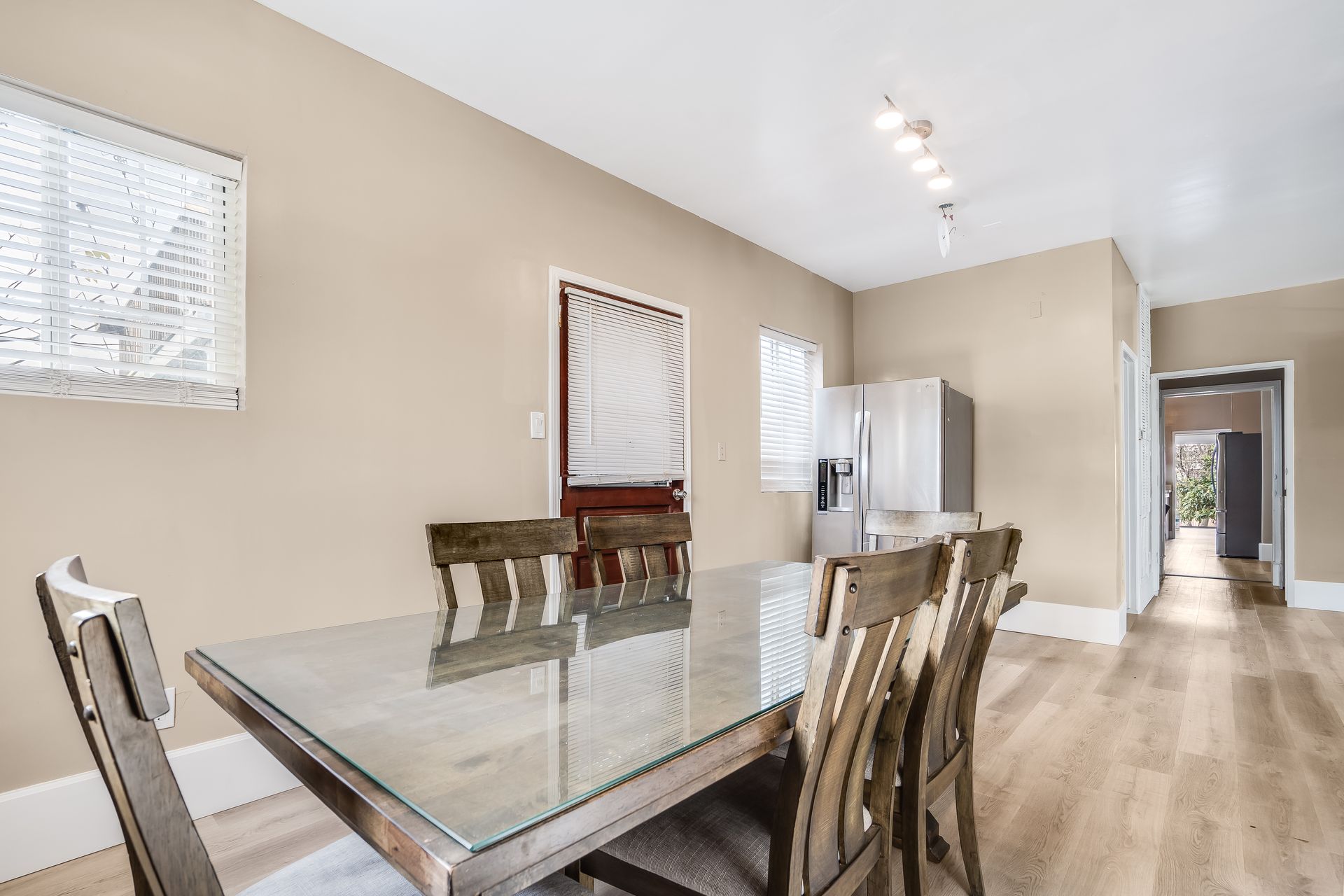 Dining room with glass-topped table, wooden chairs, beige walls, and a stainless steel refrigerator.