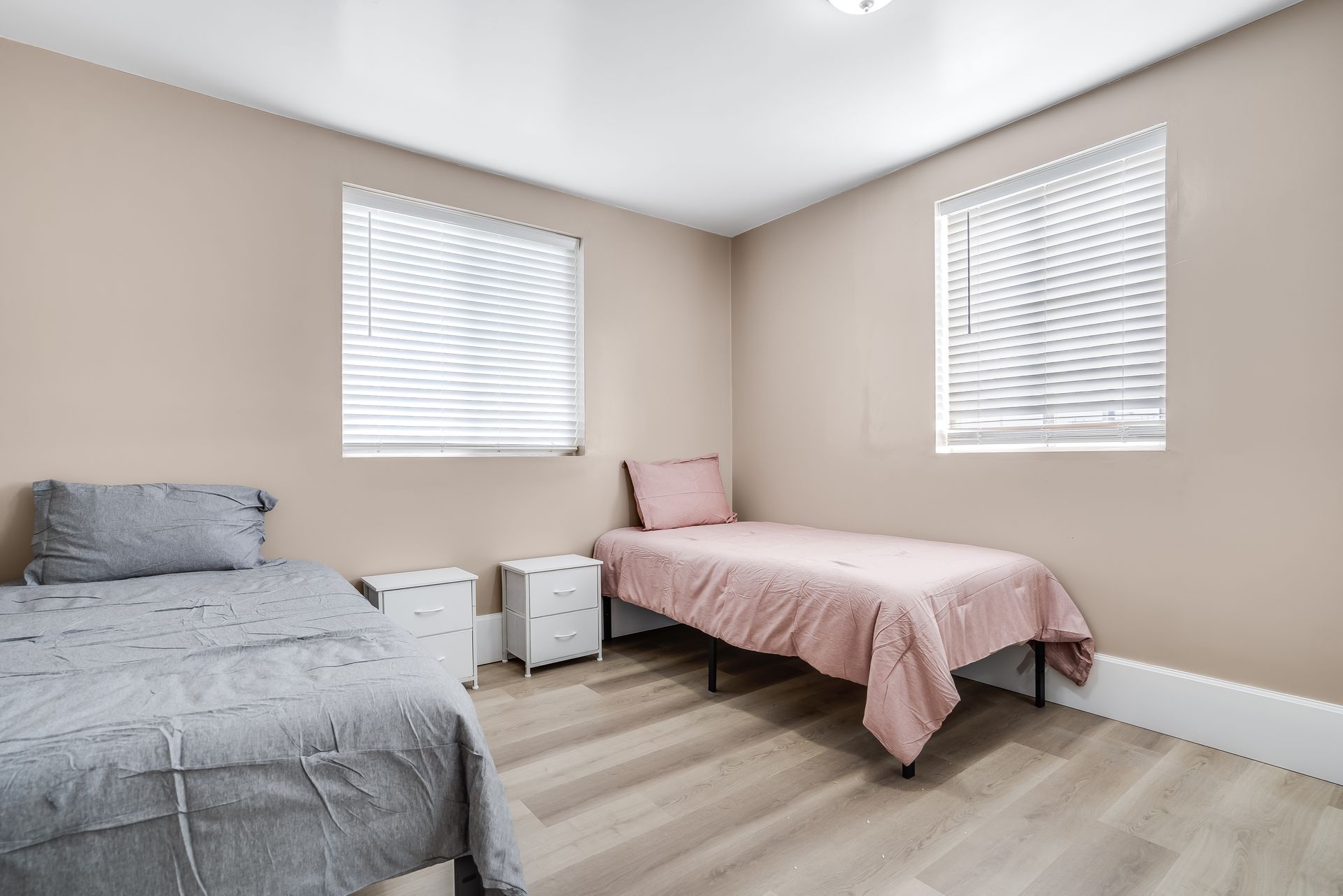 Bedroom with two twin beds, neutral walls, blinds, and wood-look flooring. One bed has gray bedding, the other pink.